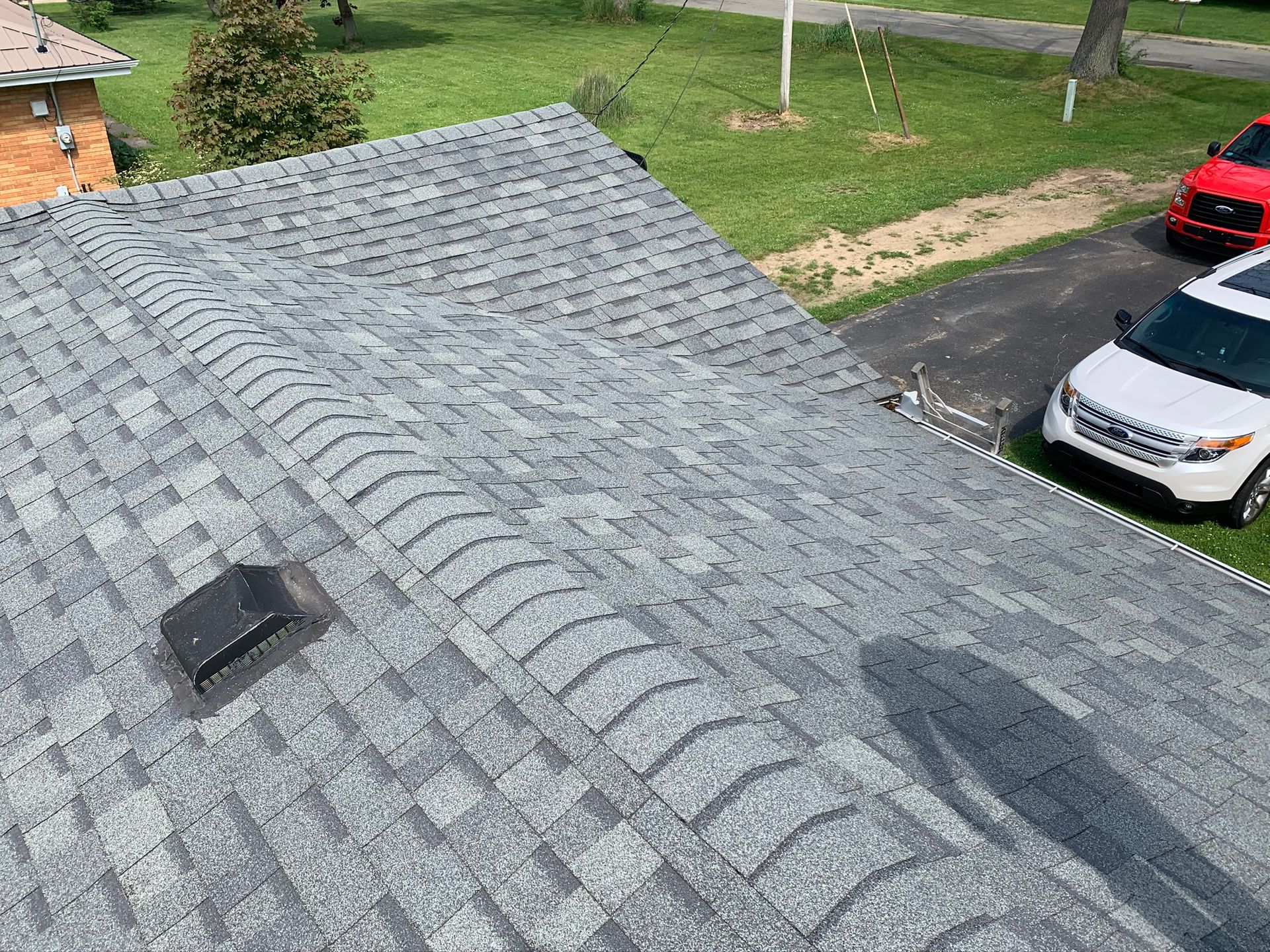 Gray shingled roof with vents, overlooking a driveway, red car, and white van below
