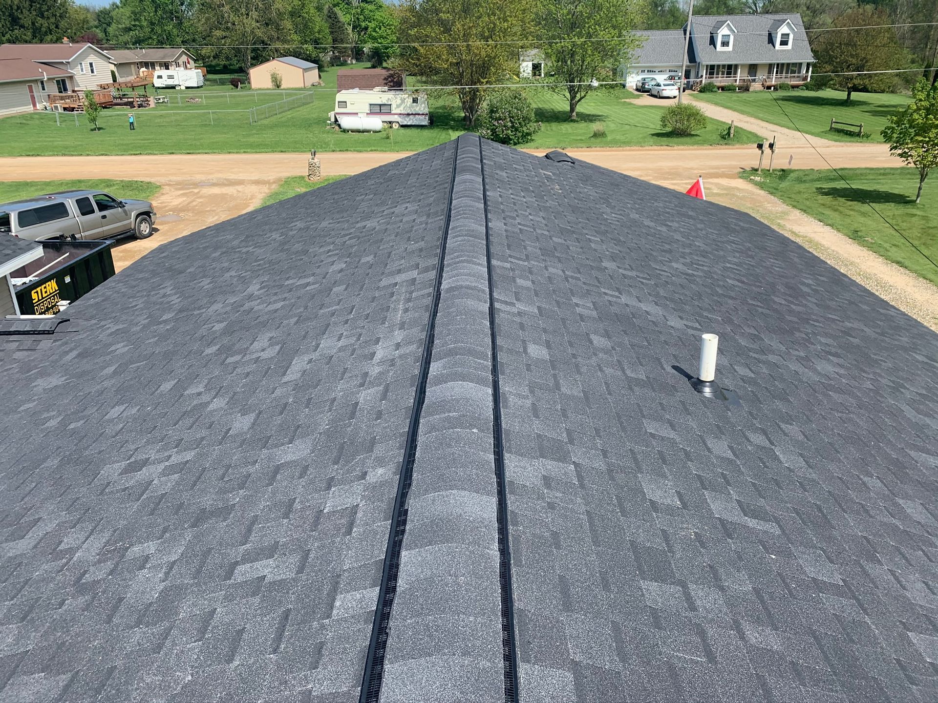 Roof shingle installation on a house, viewed from the ridge with a vent pipe and neighborhood in the background