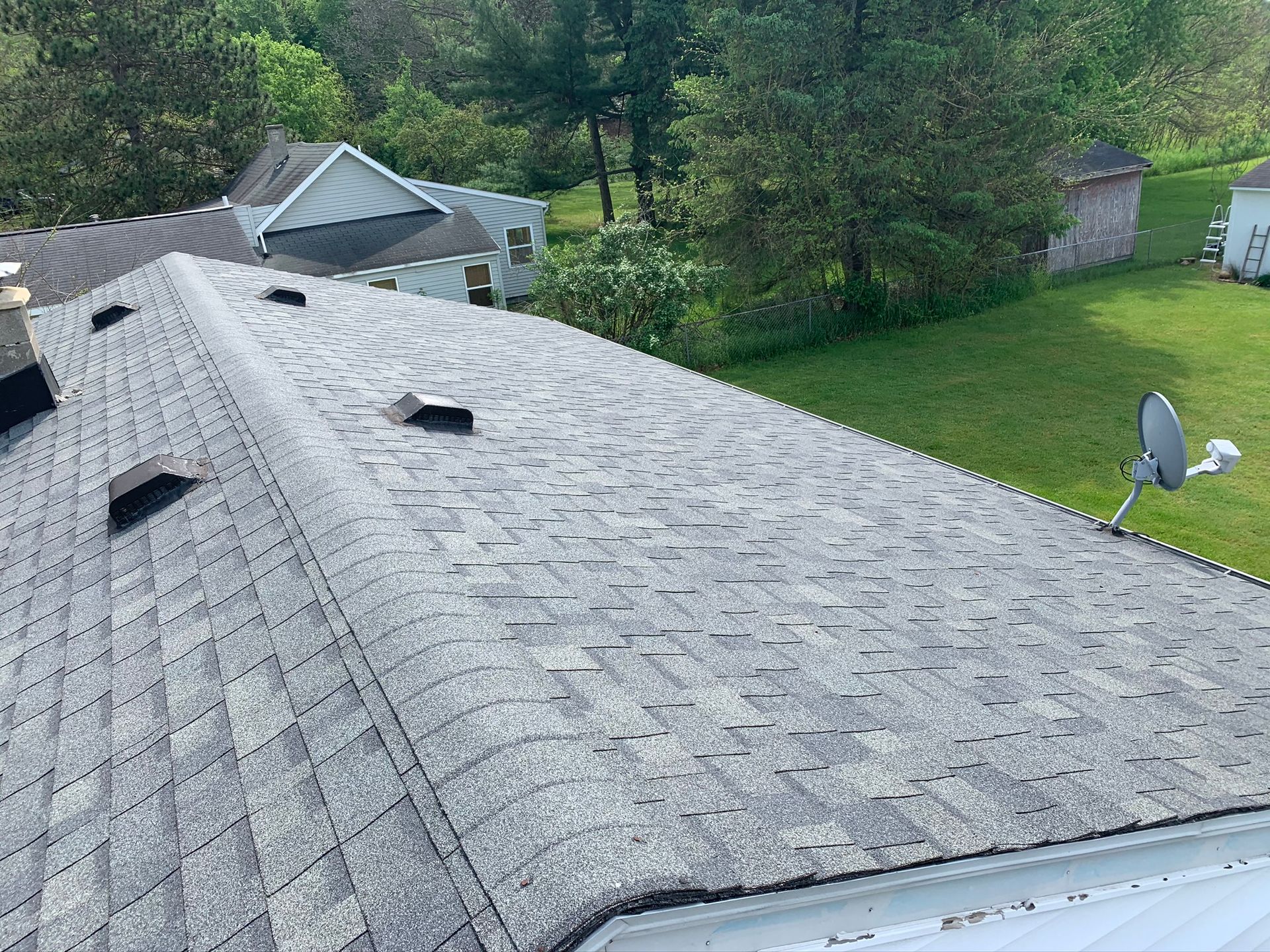 Gray shingled roof with vents overlooking a green backyard and trees