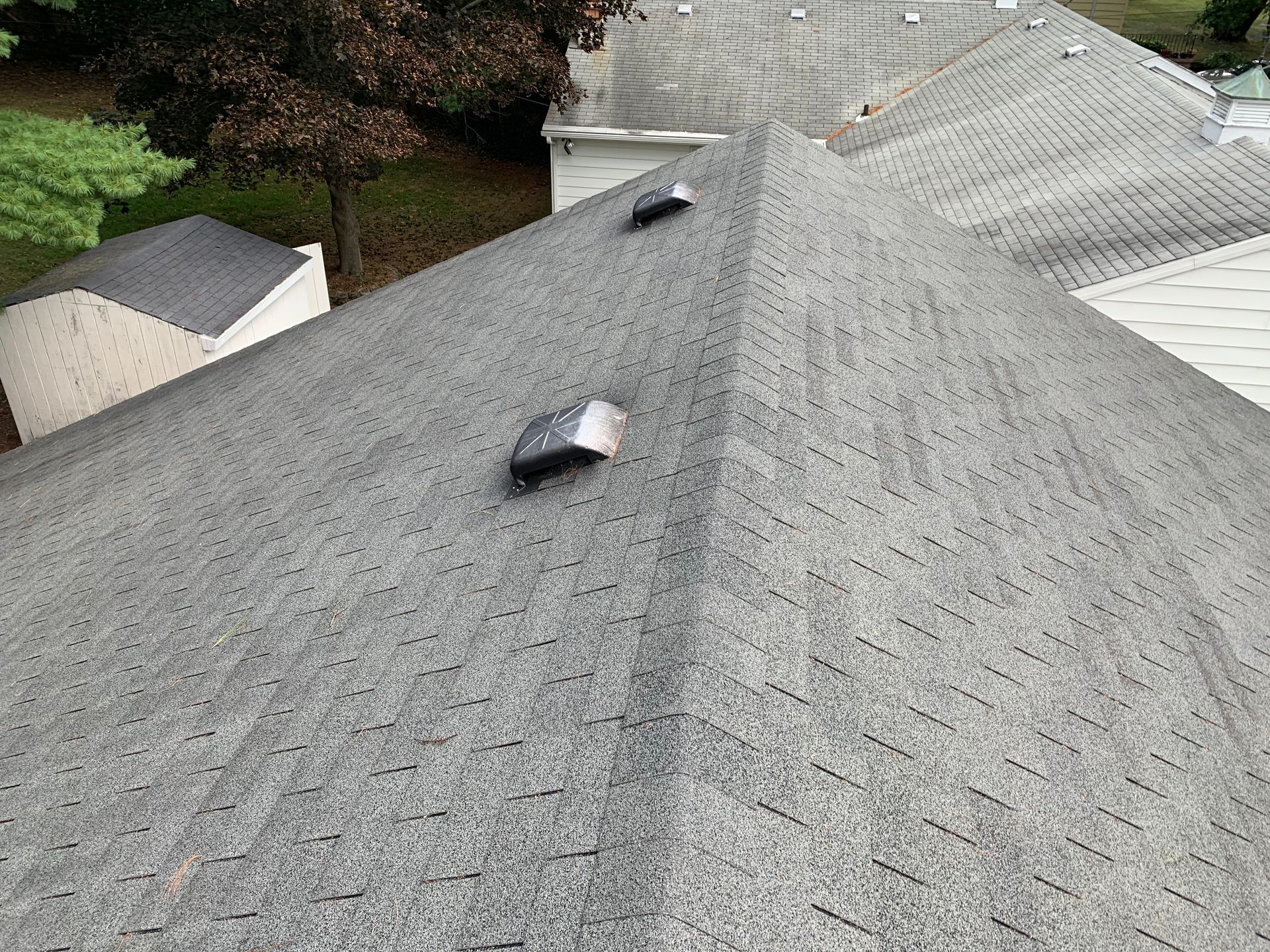 Gray shingled roof with two small vents, viewed from above, with trees and neighboring roofs nearby