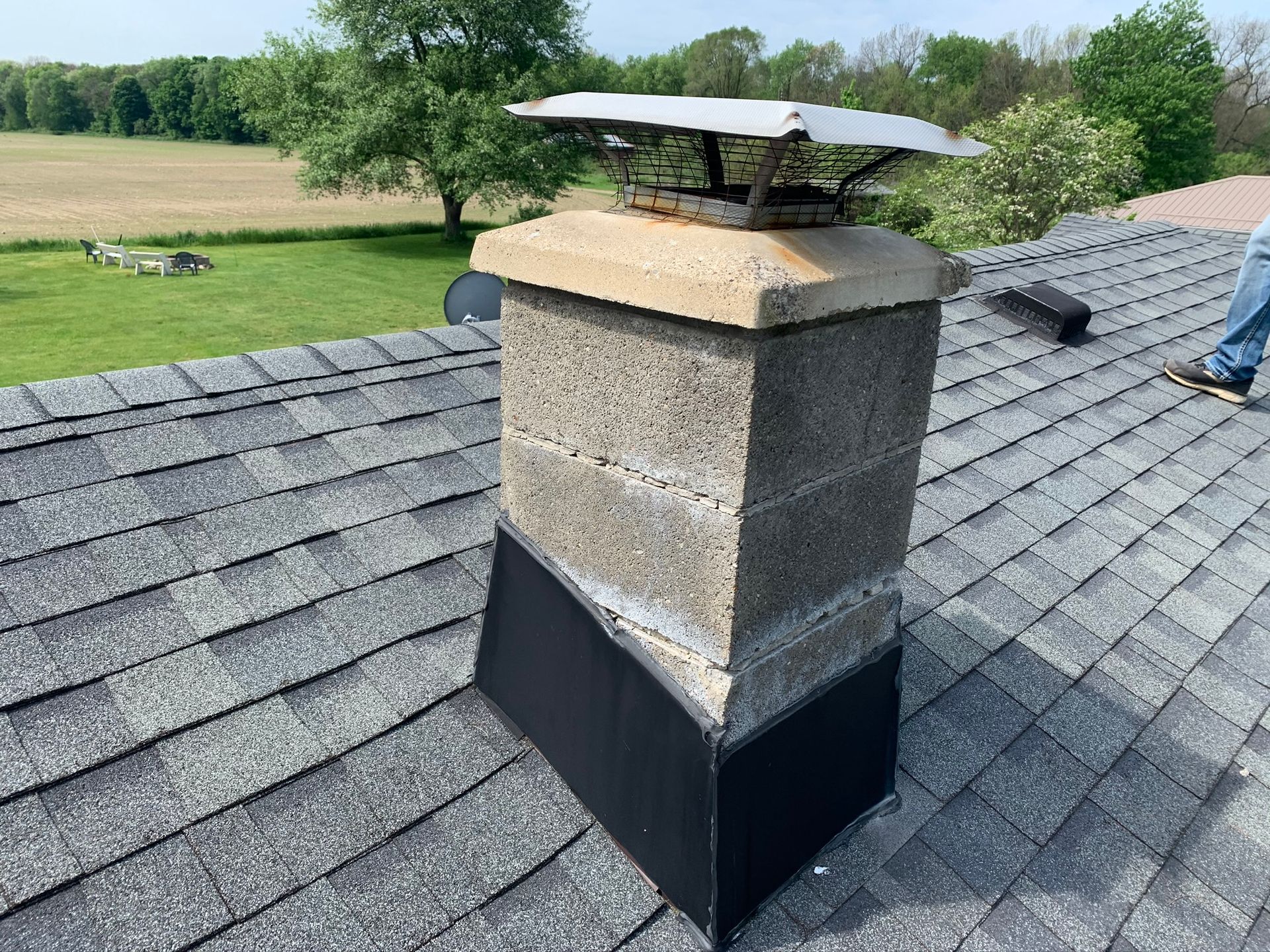 Stone chimney on a gray shingled roof with a metal cap, overlooking a green rural landscape.