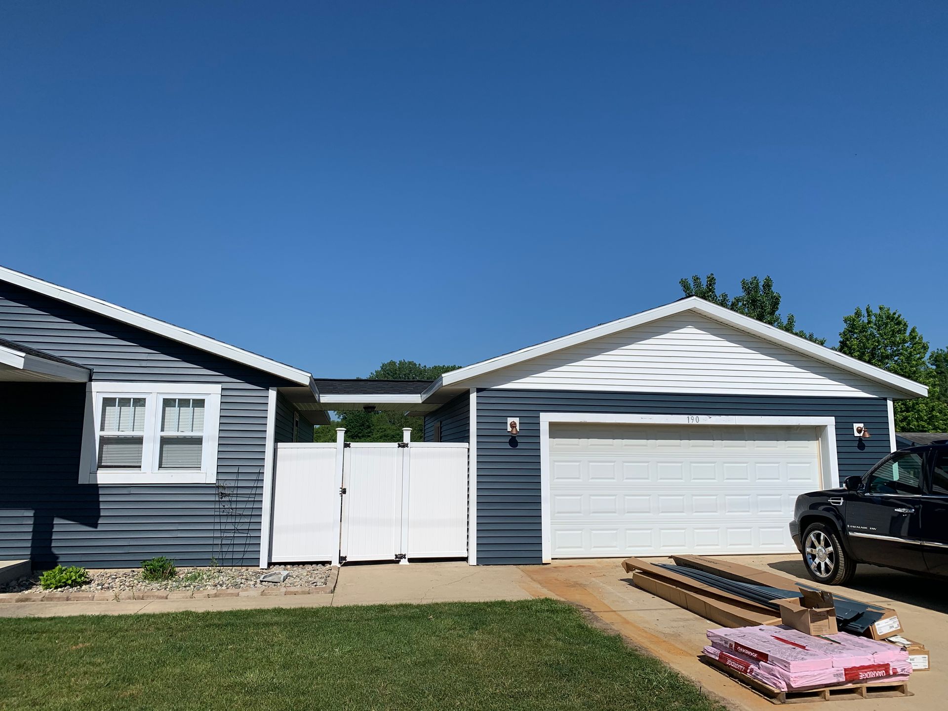 Blue house exterior with white garage door and fence, lawn in front, and stacked lumber on the driveway