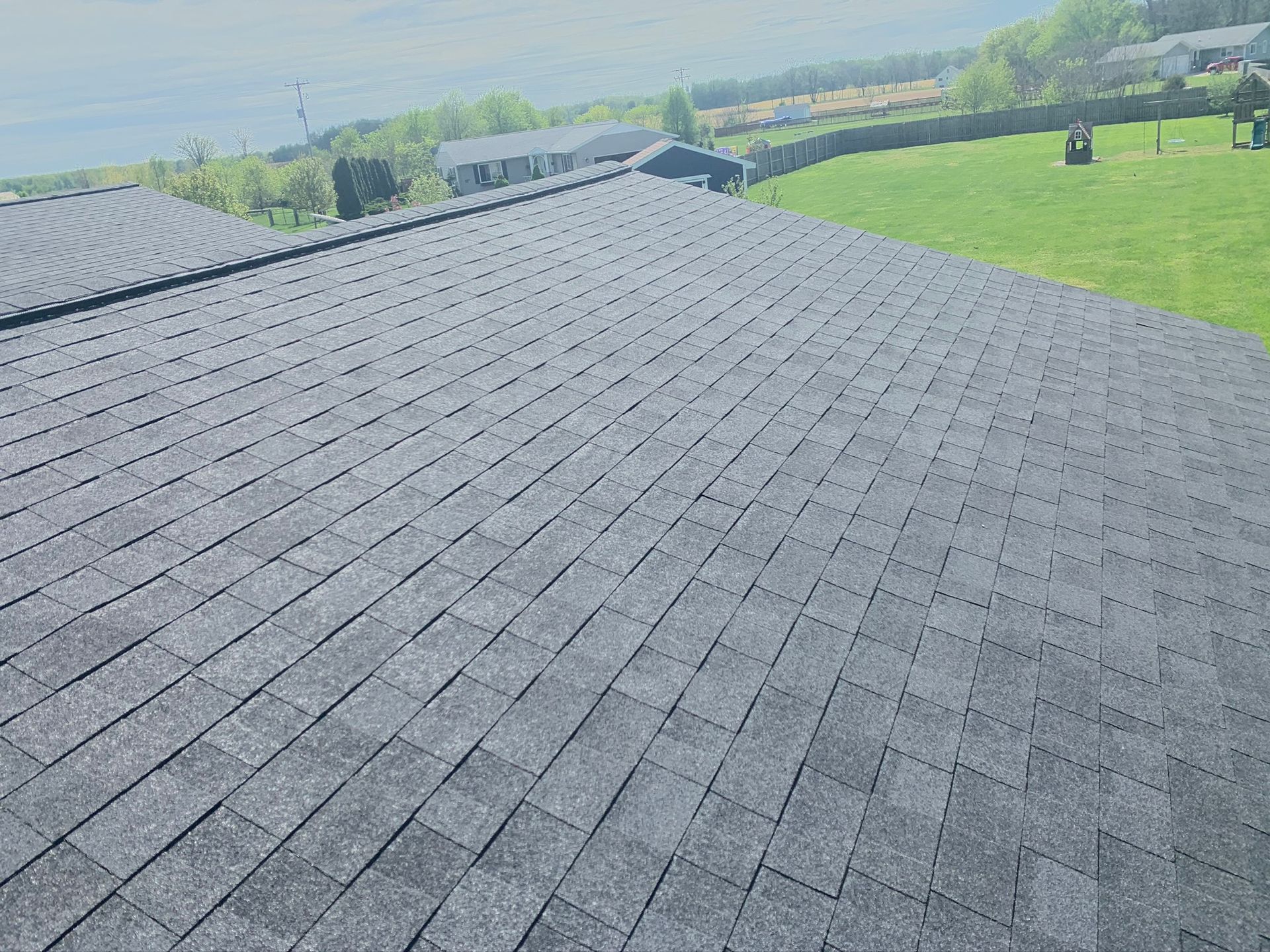 Dark gray shingled rooftop overlooking green fields and distant houses under a hazy sky