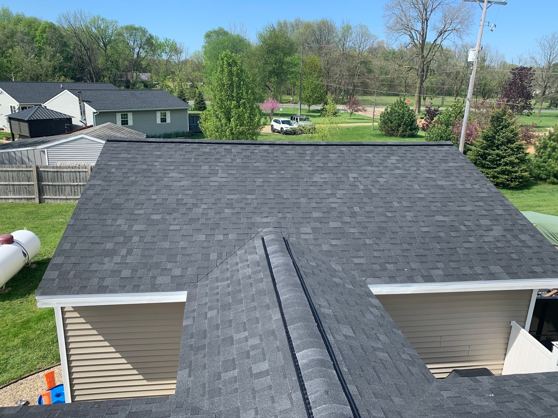 Aerial view of a gray shingled roof with two vent pipes, surrounded by trees and neighboring homes.