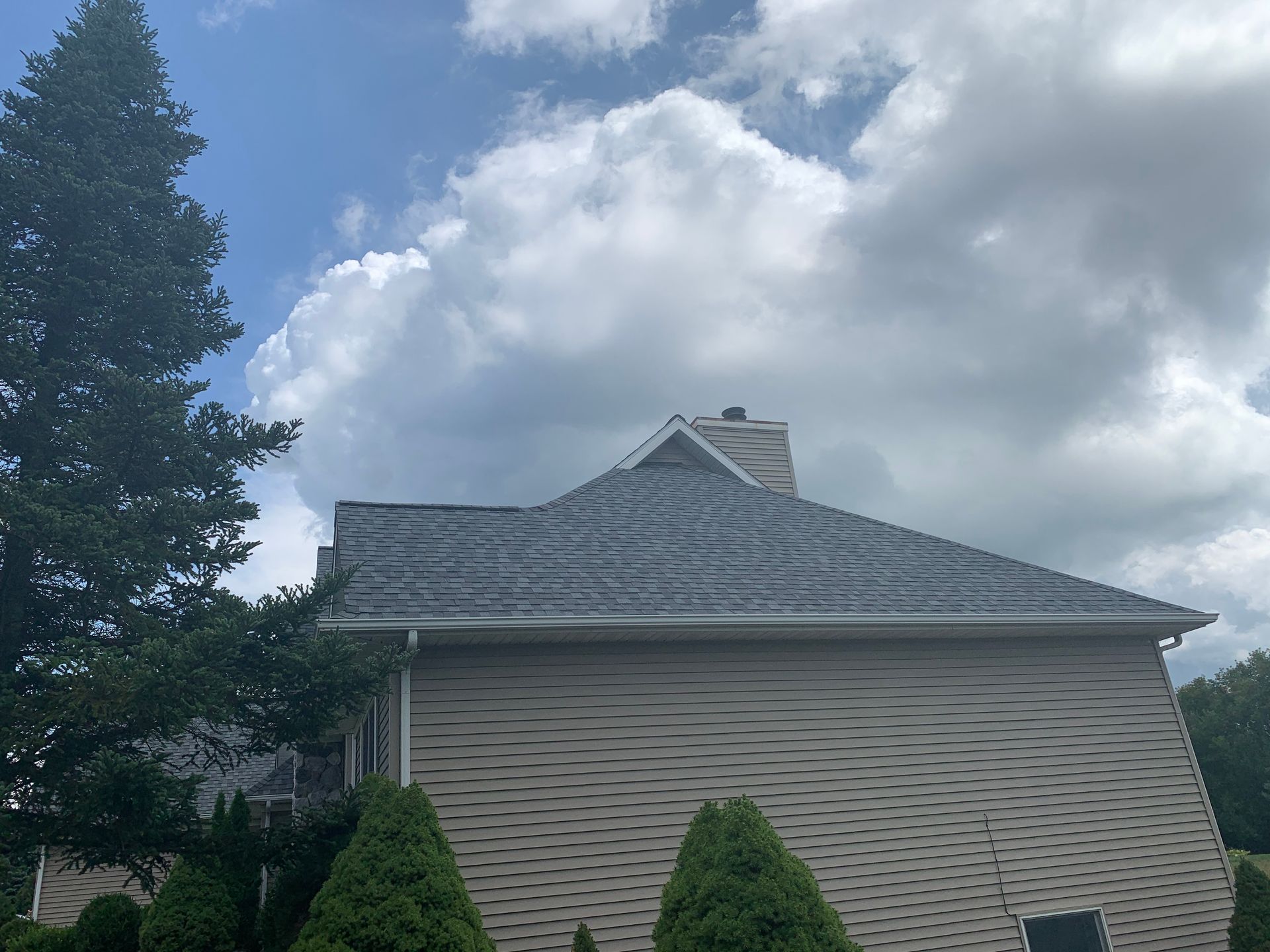 Suburban house with gray roof and beige siding under a cloudy sky, partly framed by a tall tree