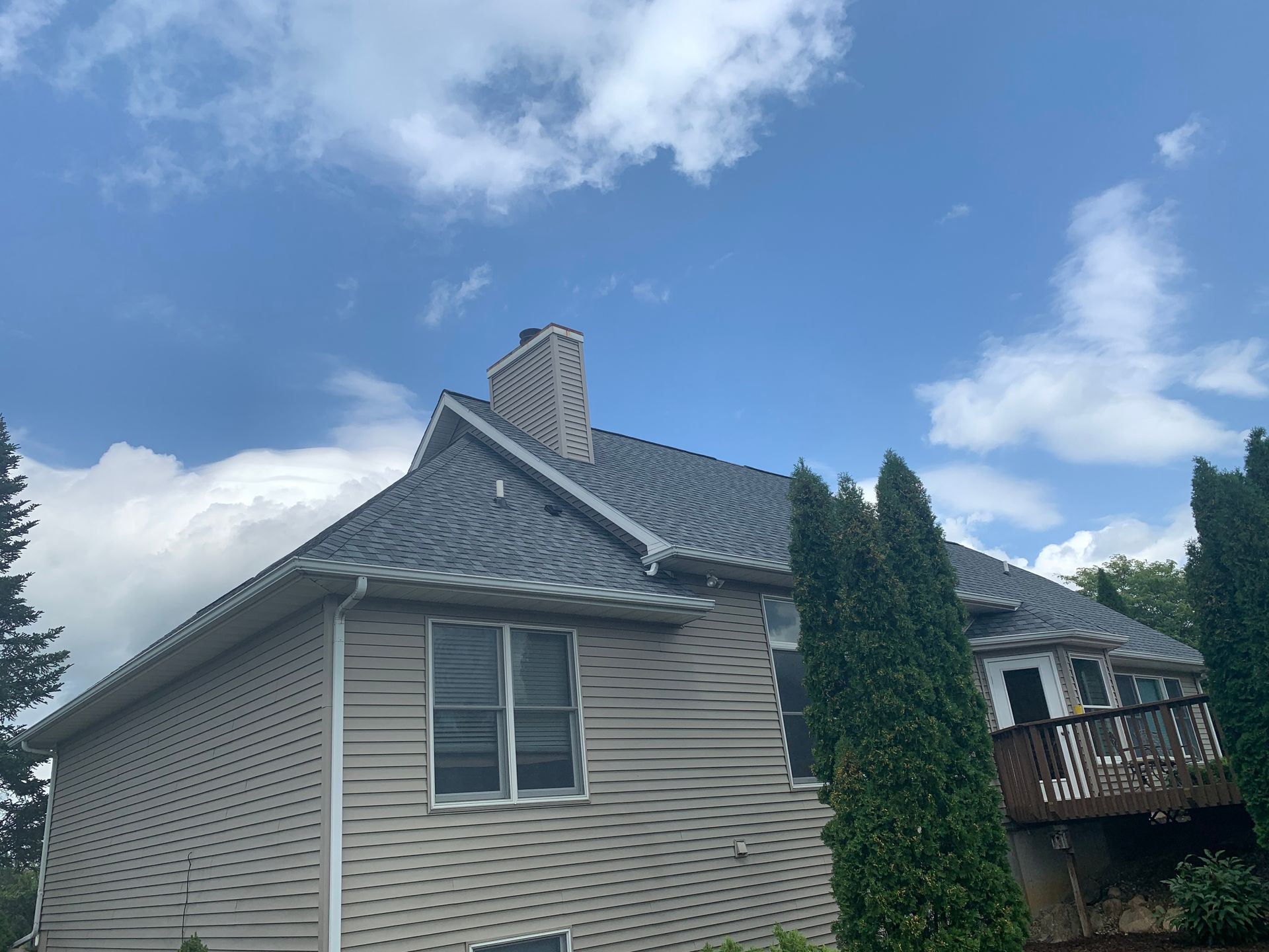 Suburban house with gray siding, blue roof, and tall evergreen trees under a partly cloudy sky