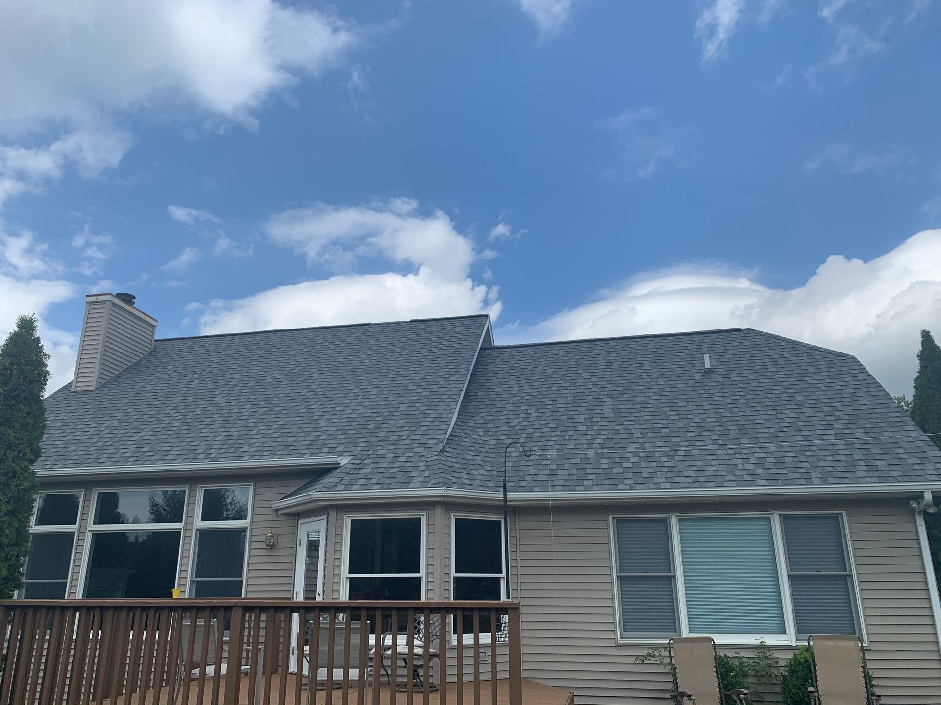 House with a gray shingled roof, large windows, and a wooden deck under a blue sky with clouds