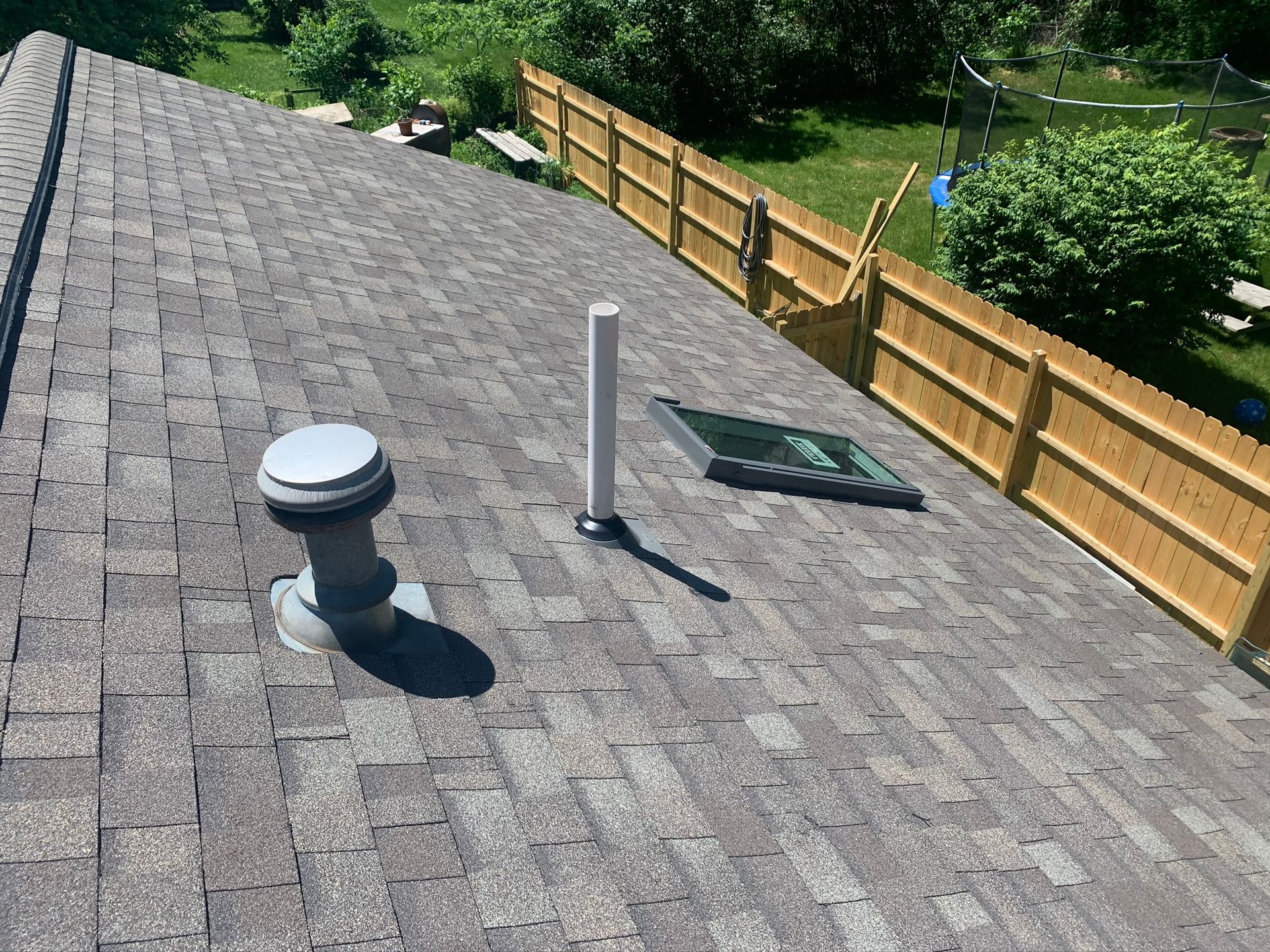 Roof with gray shingles, vent pipes, and a skylight beside a wooden fence and green trees.