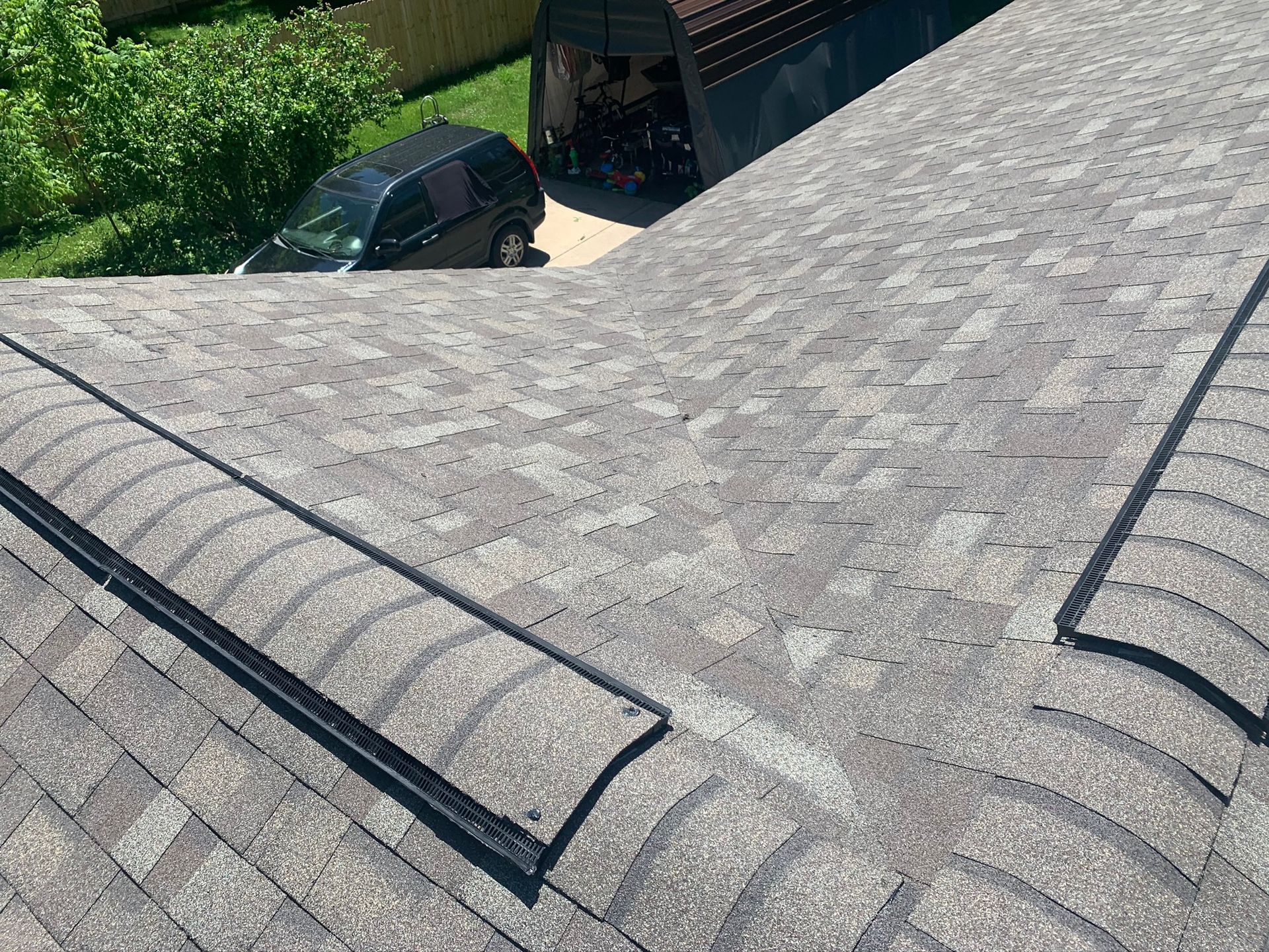 Gray shingled roof with two black roof vents, viewed from above, with trees and a parked car below