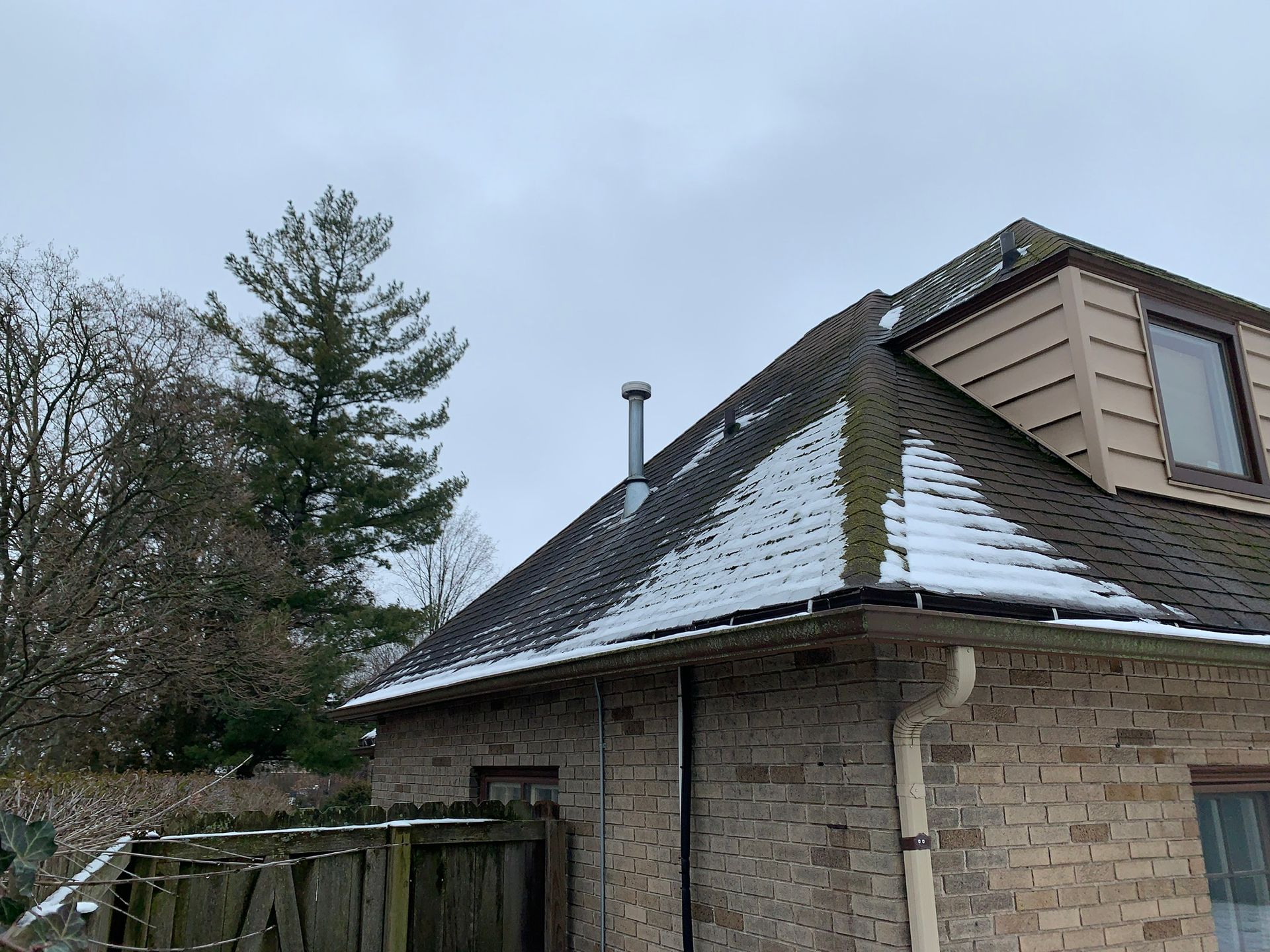 Snow-covered roof on a brick house with a wooden fence and trees nearby