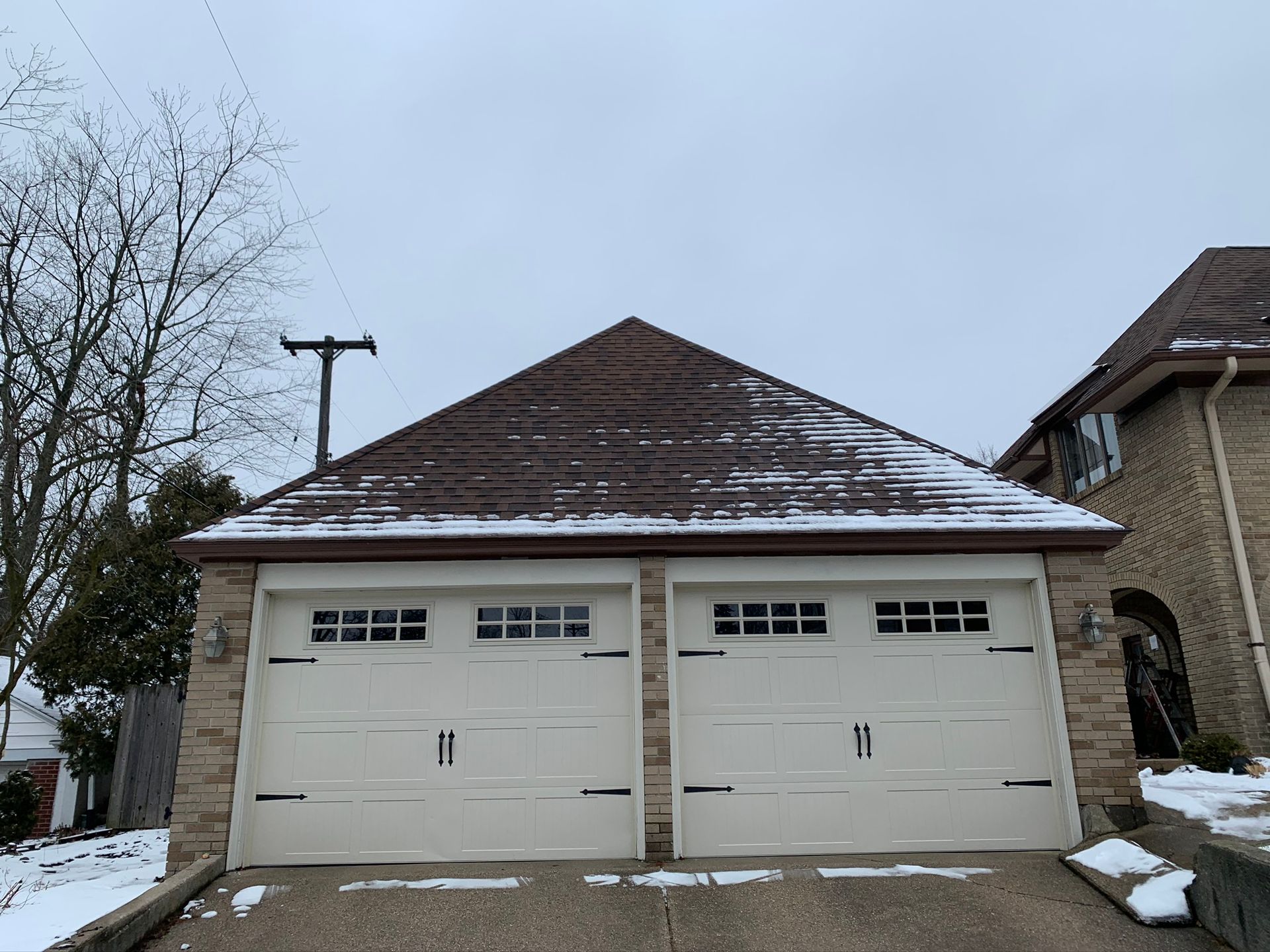 Snow-dusted detached garage with three white doors and a brown shingled roof beside a house.