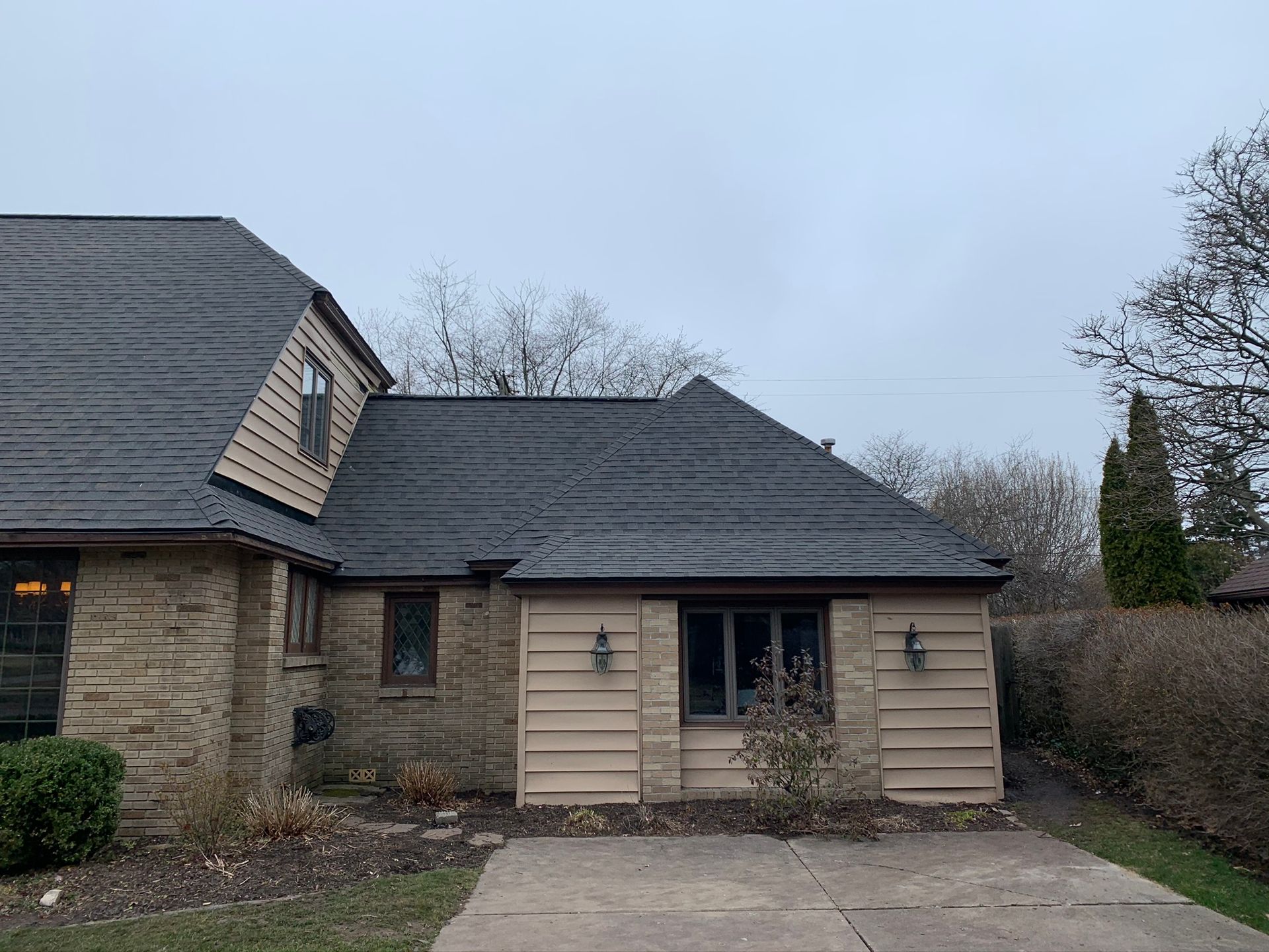 Stone house exterior with dark shingled roof, tan siding, and a small front porch on an overcast day