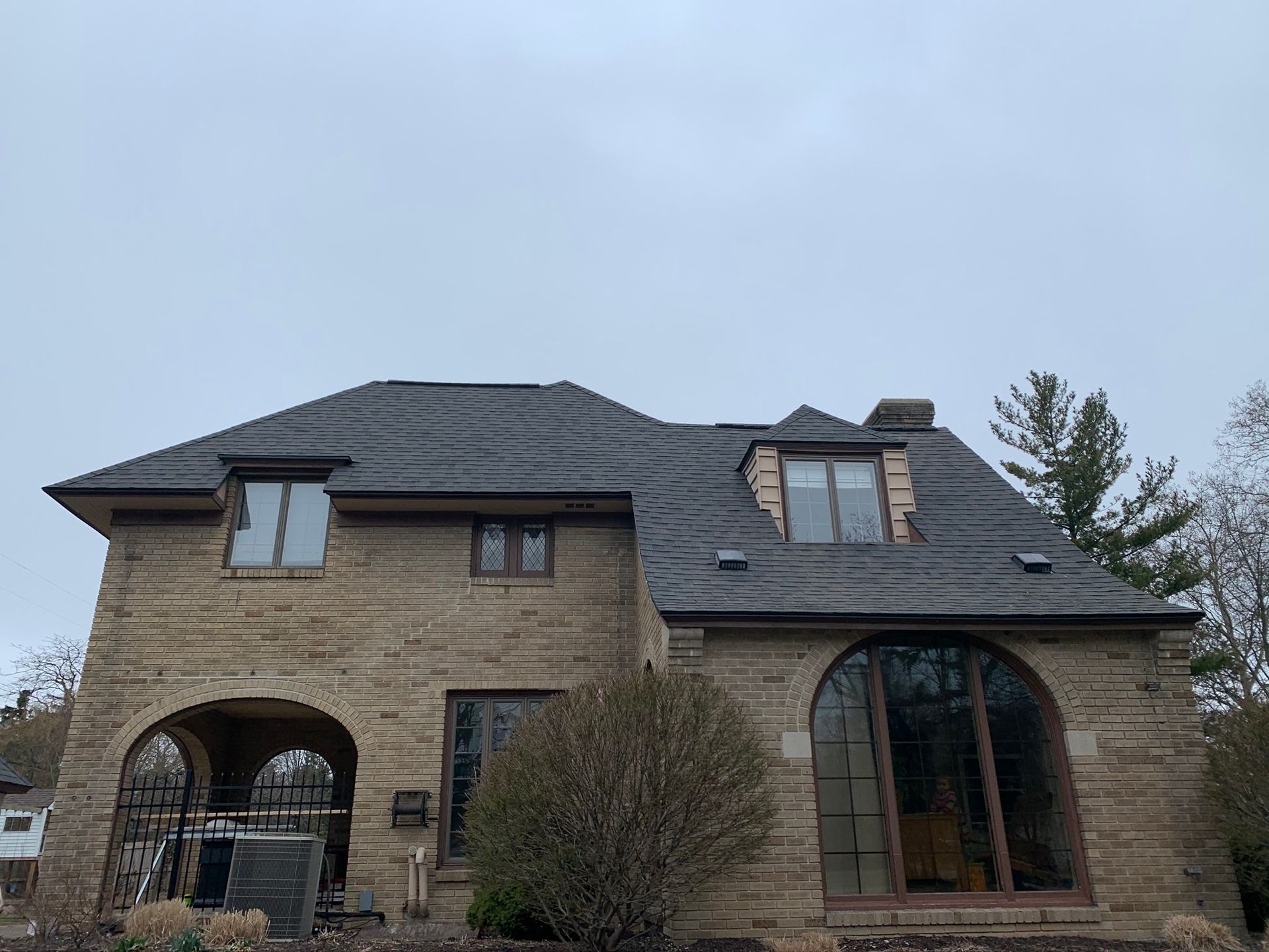 Two-story stone house with dark roof, arched windows, and a front shrub under an overcast sky