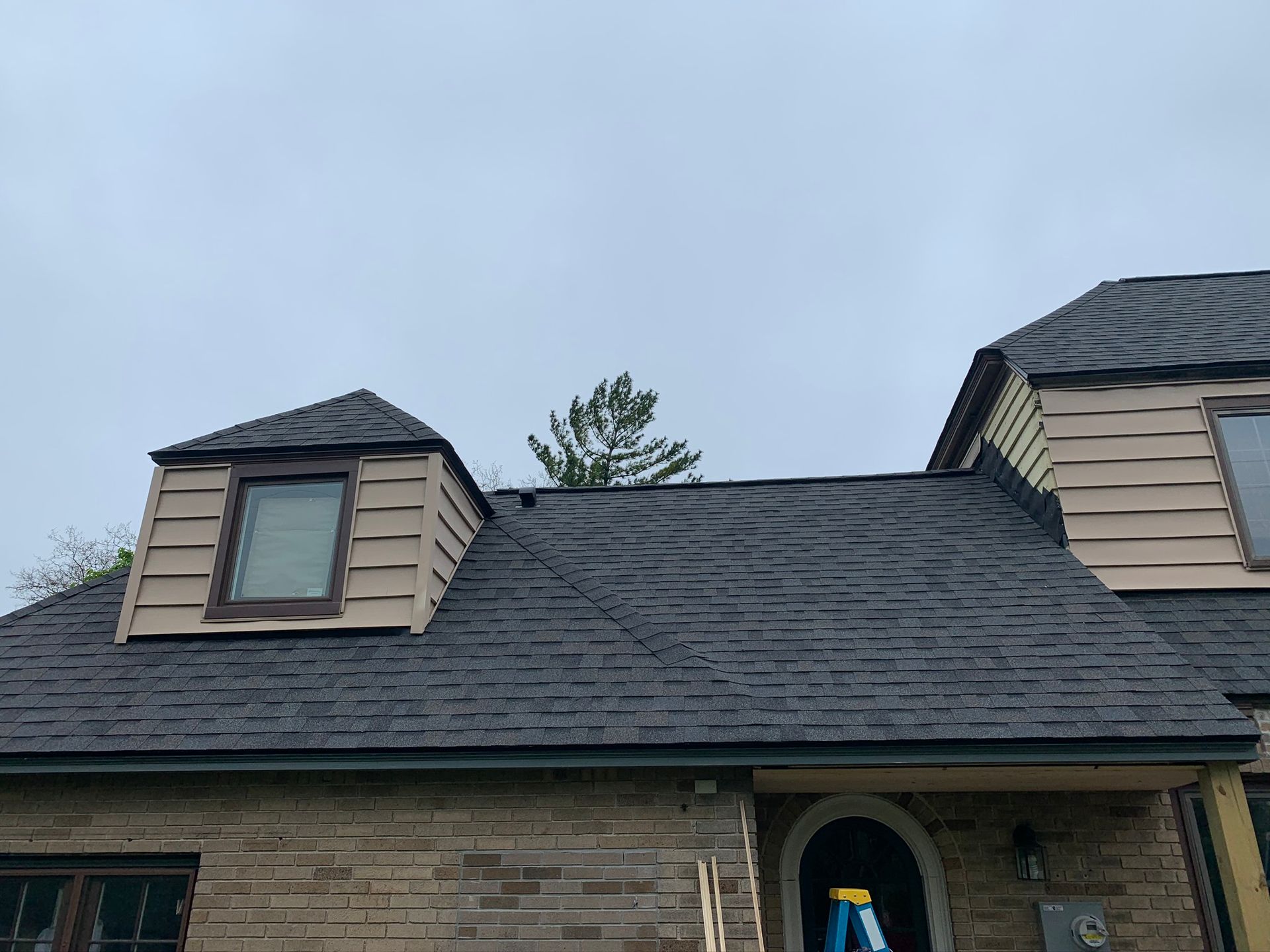 Suburban house roof with dark shingles and two dormer windows under a cloudy sky