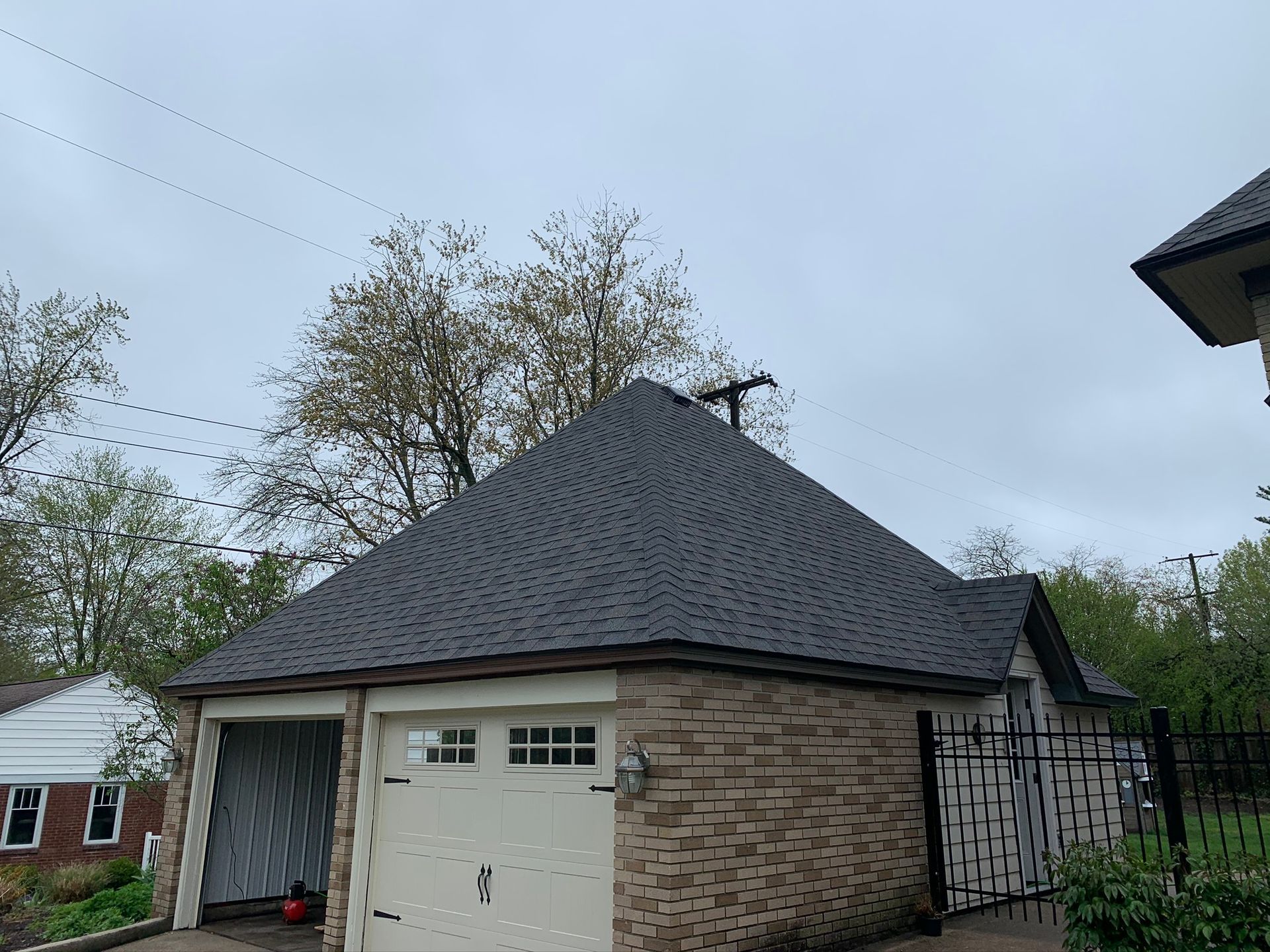 Brick garage with dark shingled roof and white door beside a fenced yard under a gray sky