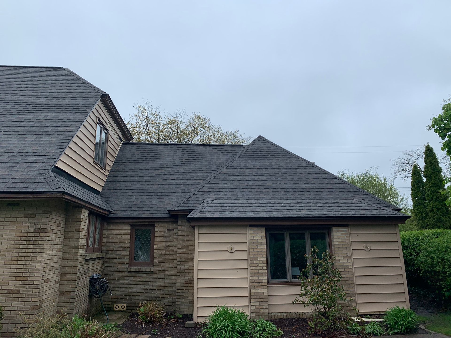 Stone-and-beige house exterior with dark gray shingled roof and small windows under an overcast sky