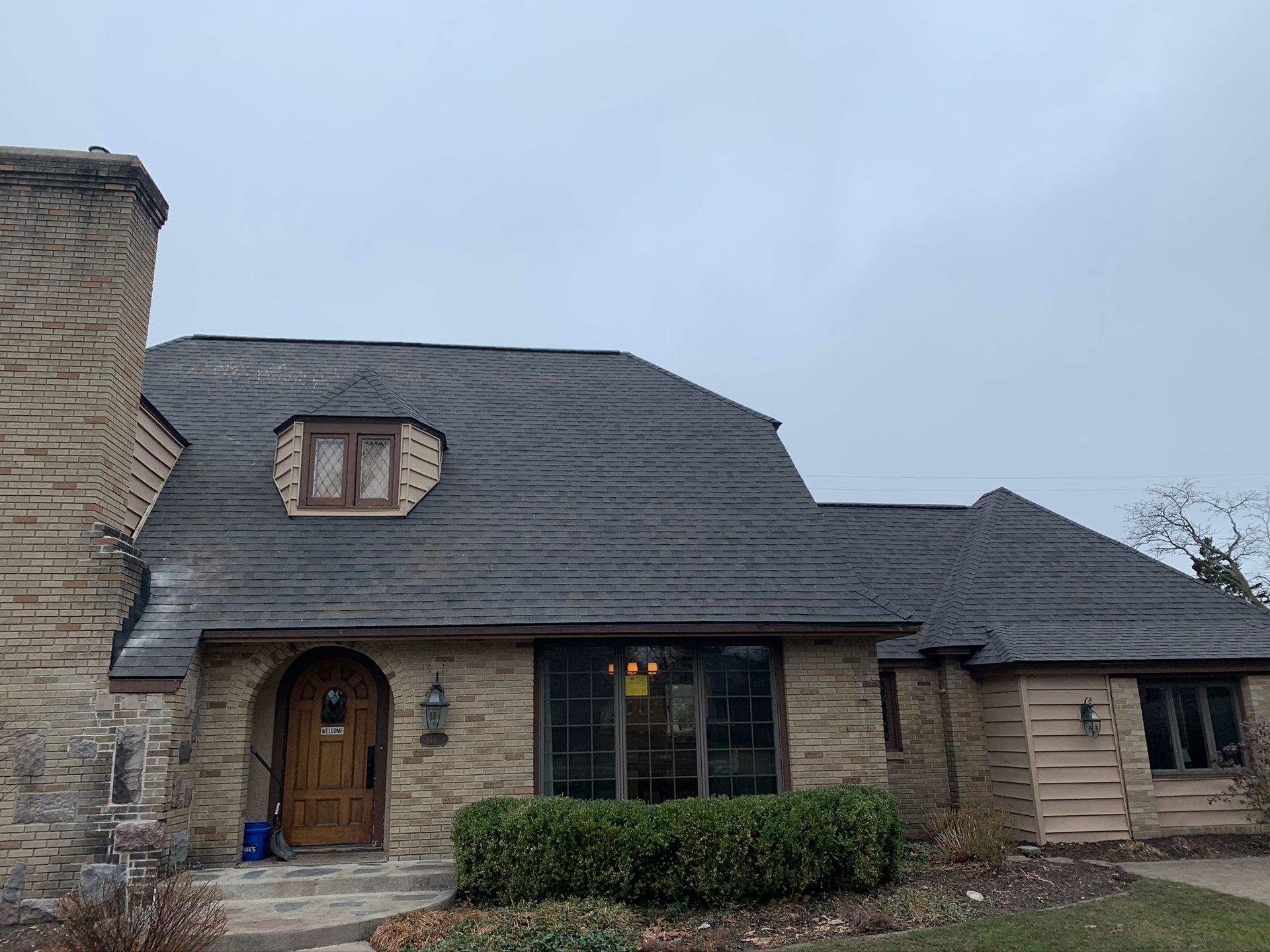 Stone house with dark gray roof, arched wooden door, and front hedge on a cloudy day