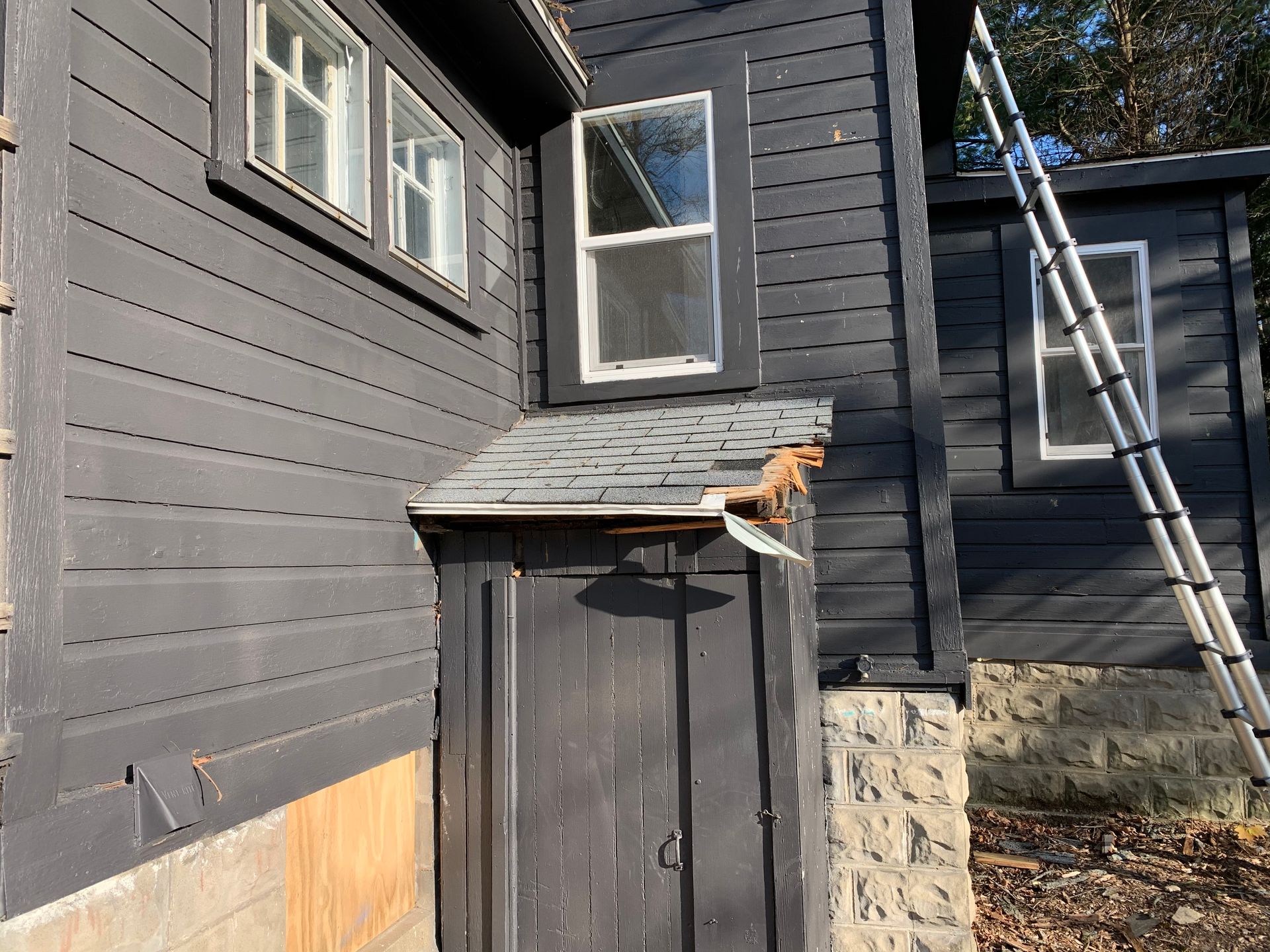 Back of dark gray house with open window, small shed, ladder, and stone foundation