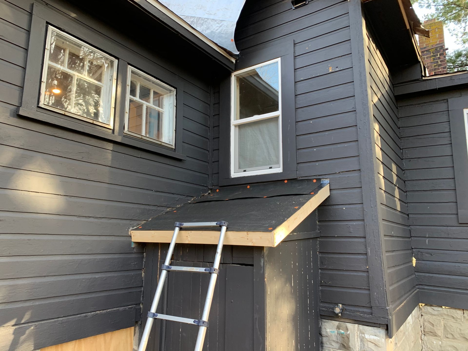 Gray house exterior with a ladder leaning against a small roof overhang and windows nearby