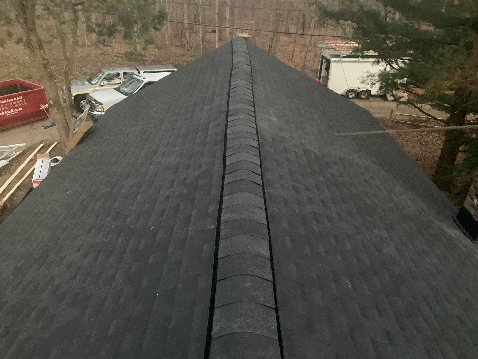 Roof ridge with dark shingles, viewed from above, with trees and vehicles in the background