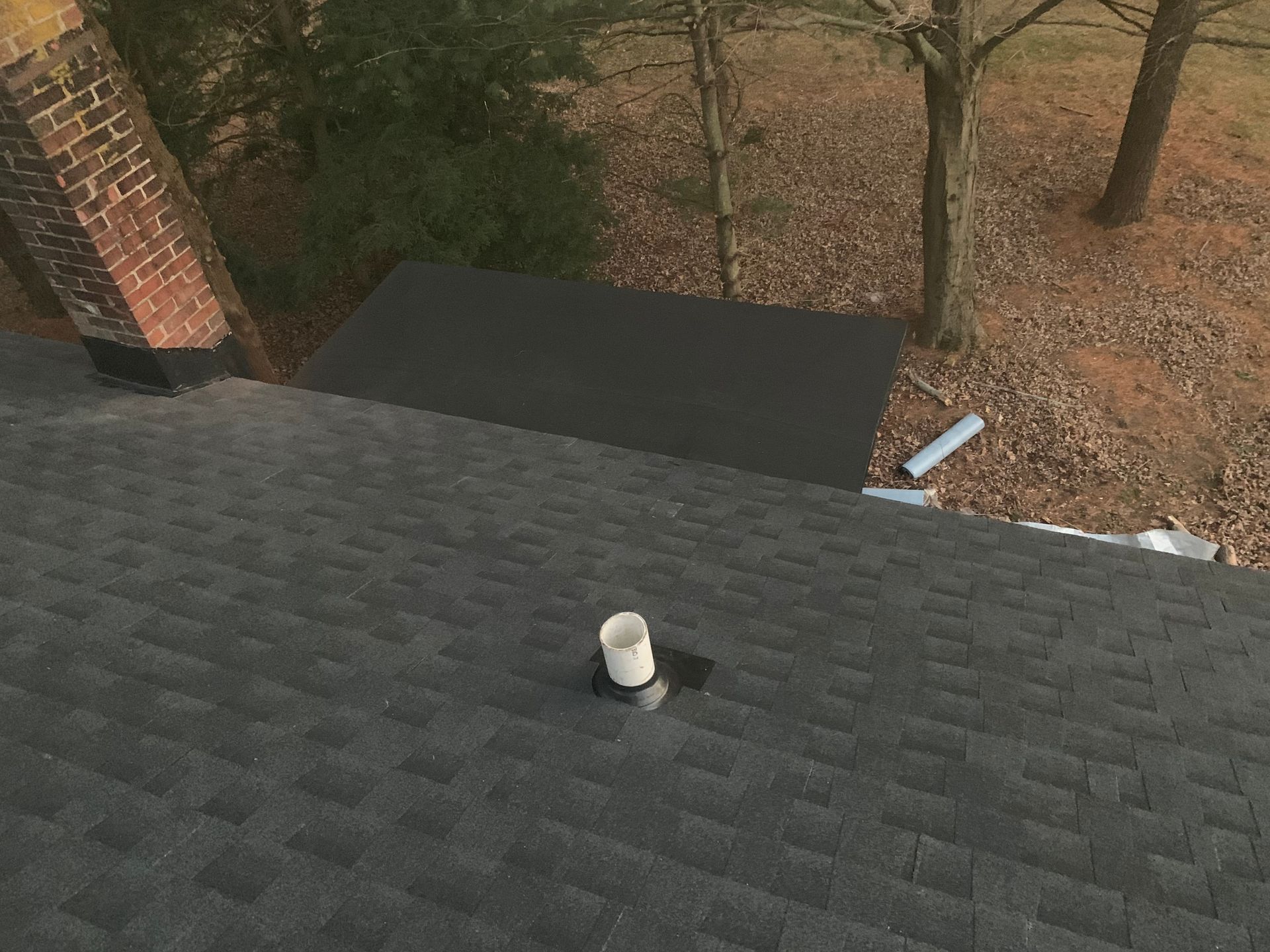 Dark shingled roof with a white vent, seen from above beside trees and a brick chimney.