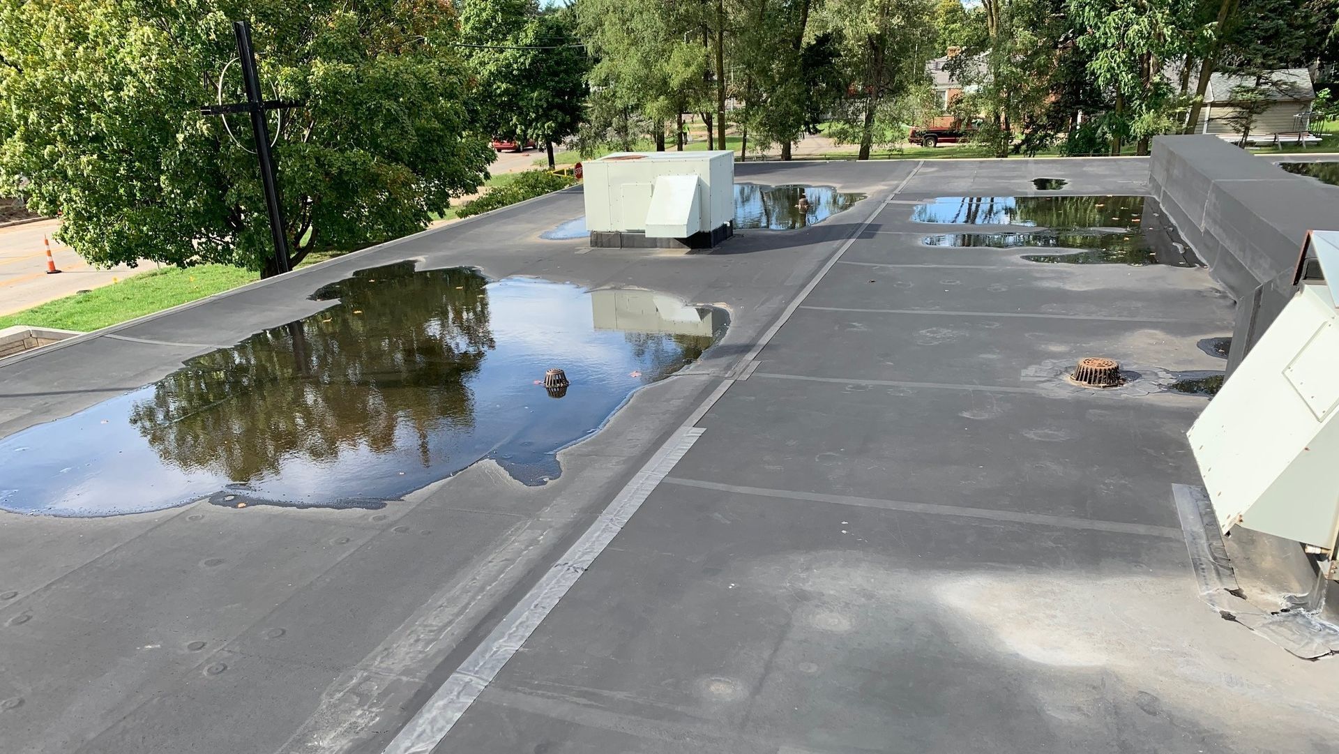 Flat rooftop with large puddles of standing water and white HVAC units near a parapet wall.