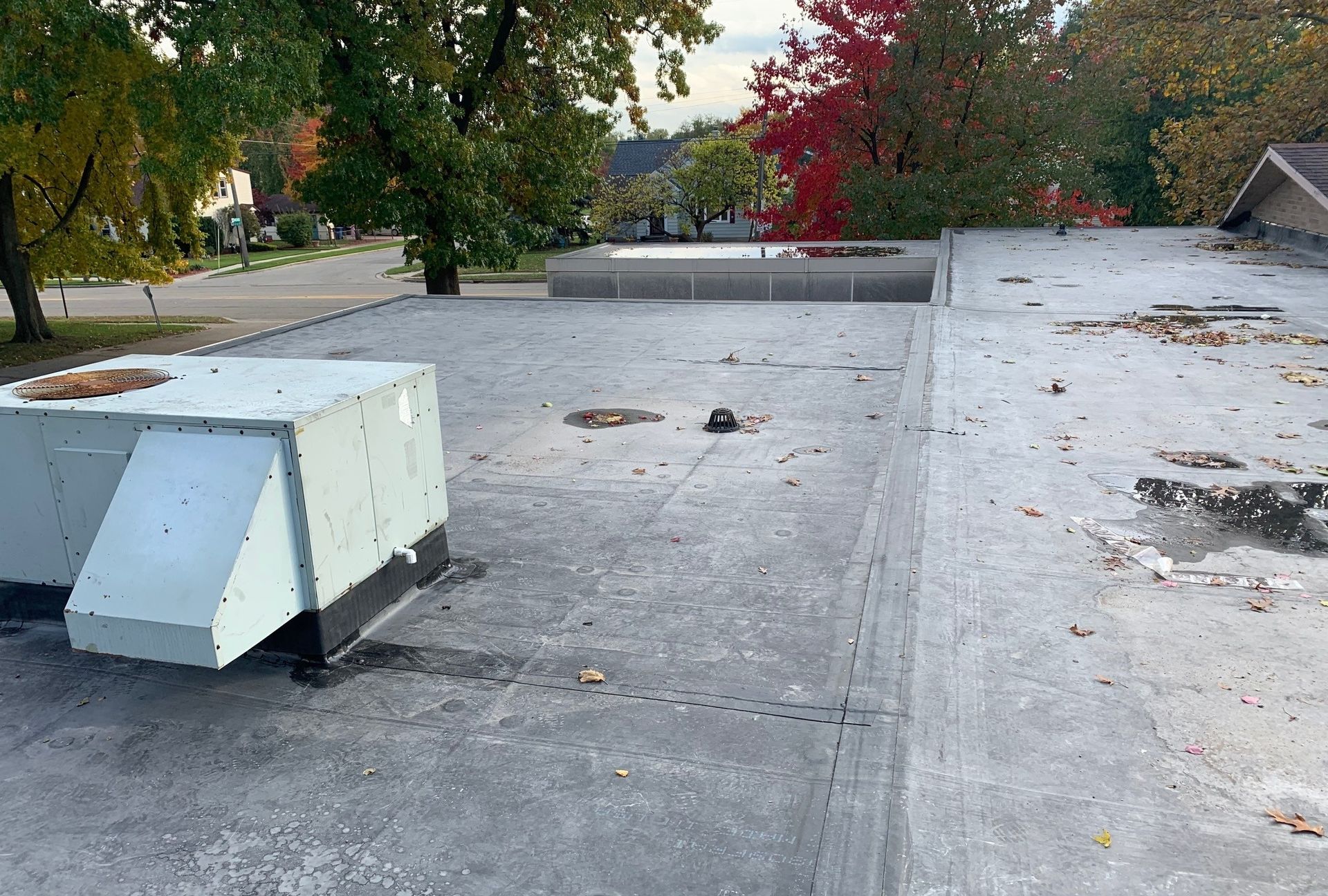 Flat rooftop with a white HVAC unit and scattered debris, trees and buildings in the background.