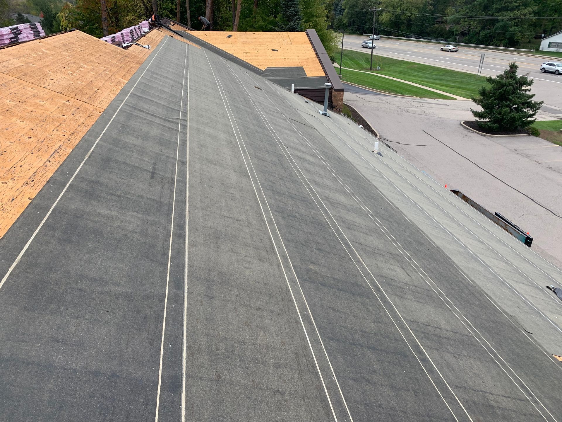 Roof under construction with gray underlayment, orange sheathing, and a parking lot below.