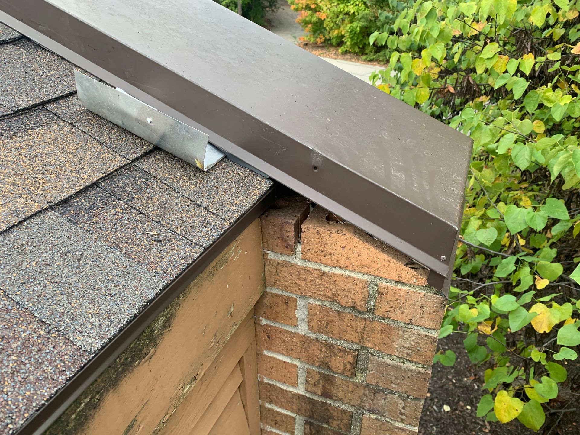 Brown metal roof edge over shingles and a brick wall beside green shrubs