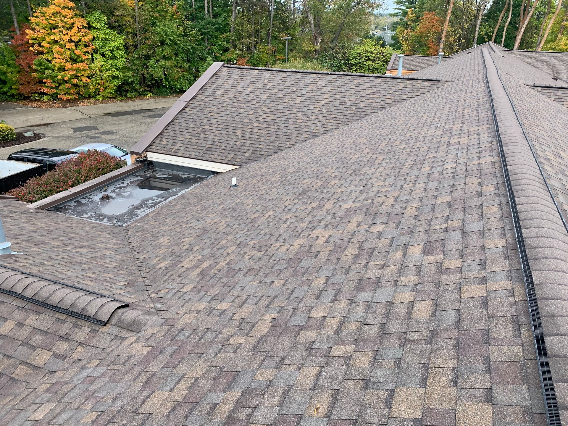 Rooftop with brown shingles and two open skylights beside trees in autumn colors