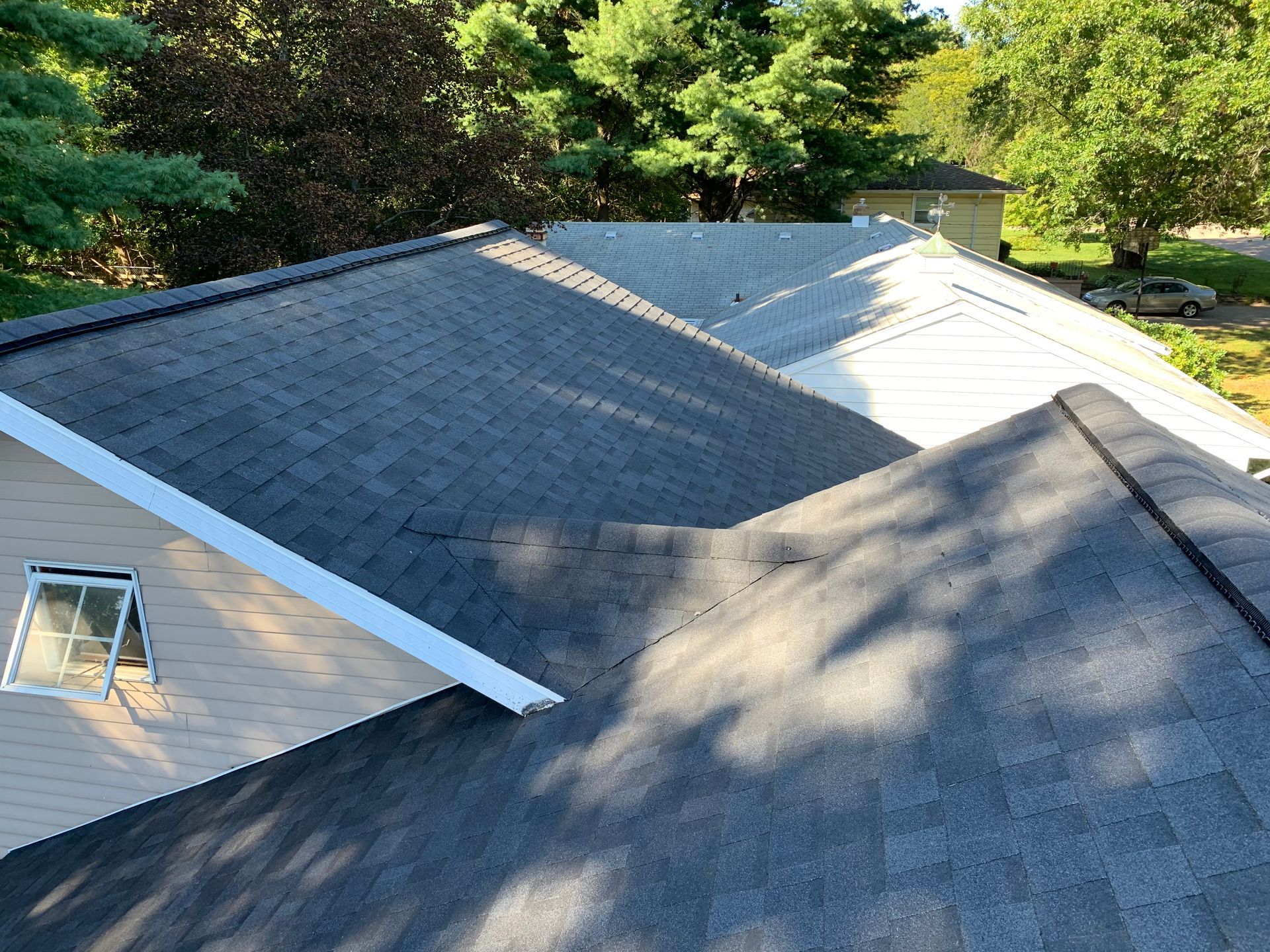 Roof with gray shingles and white trim in a wooded area, shown in bright sunlight.