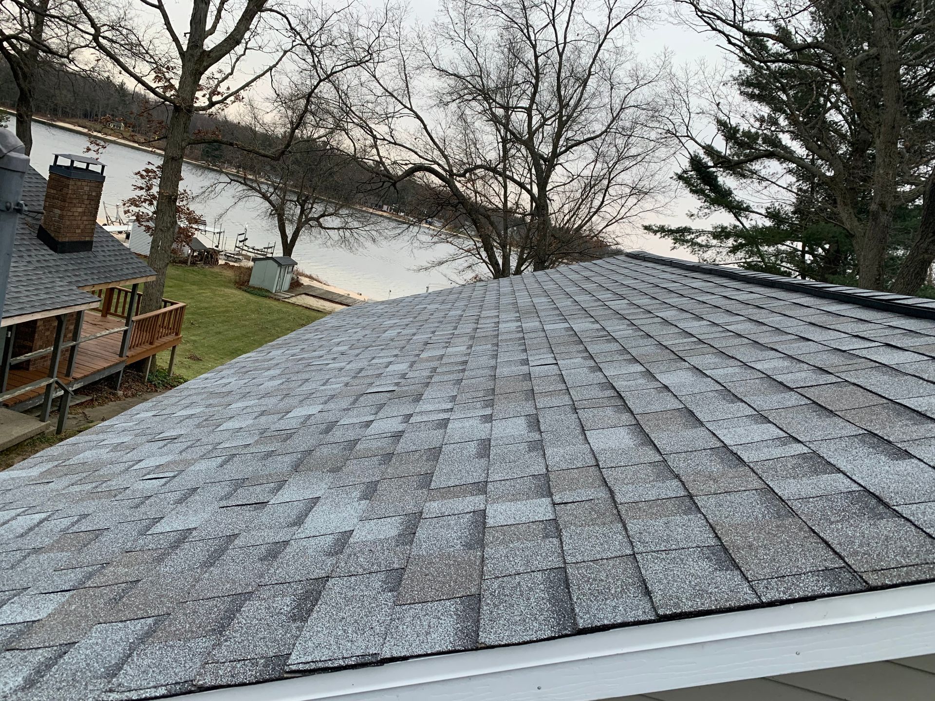 Gray shingled roof in the foreground with a yard, leafless trees, and a lake beyond