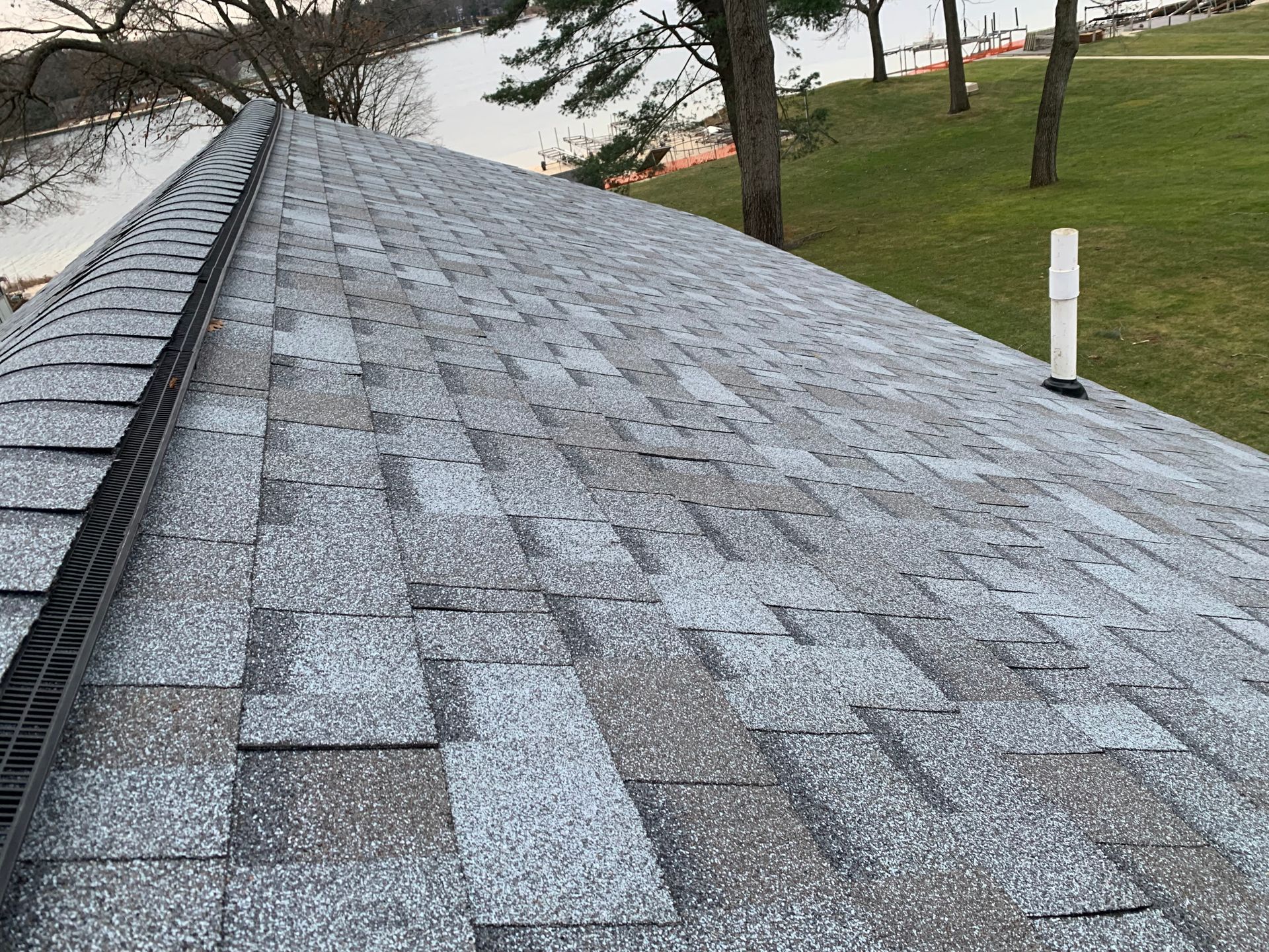 Shingled roof slope with gray shingles, a ridge vent, and trees and grass in the background