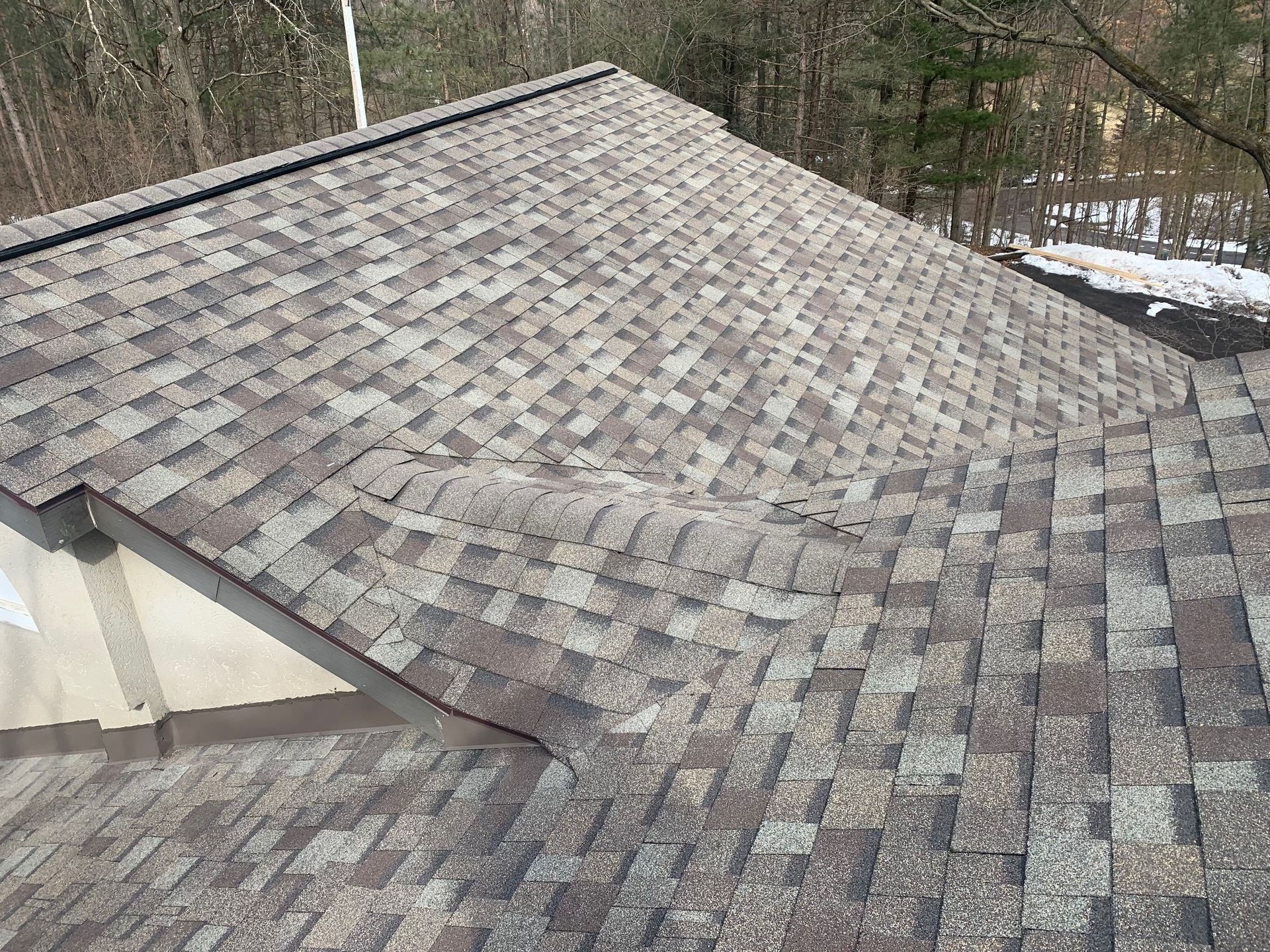 Gray shingled roof on a house with a dormer, surrounded by bare trees and patches of snow