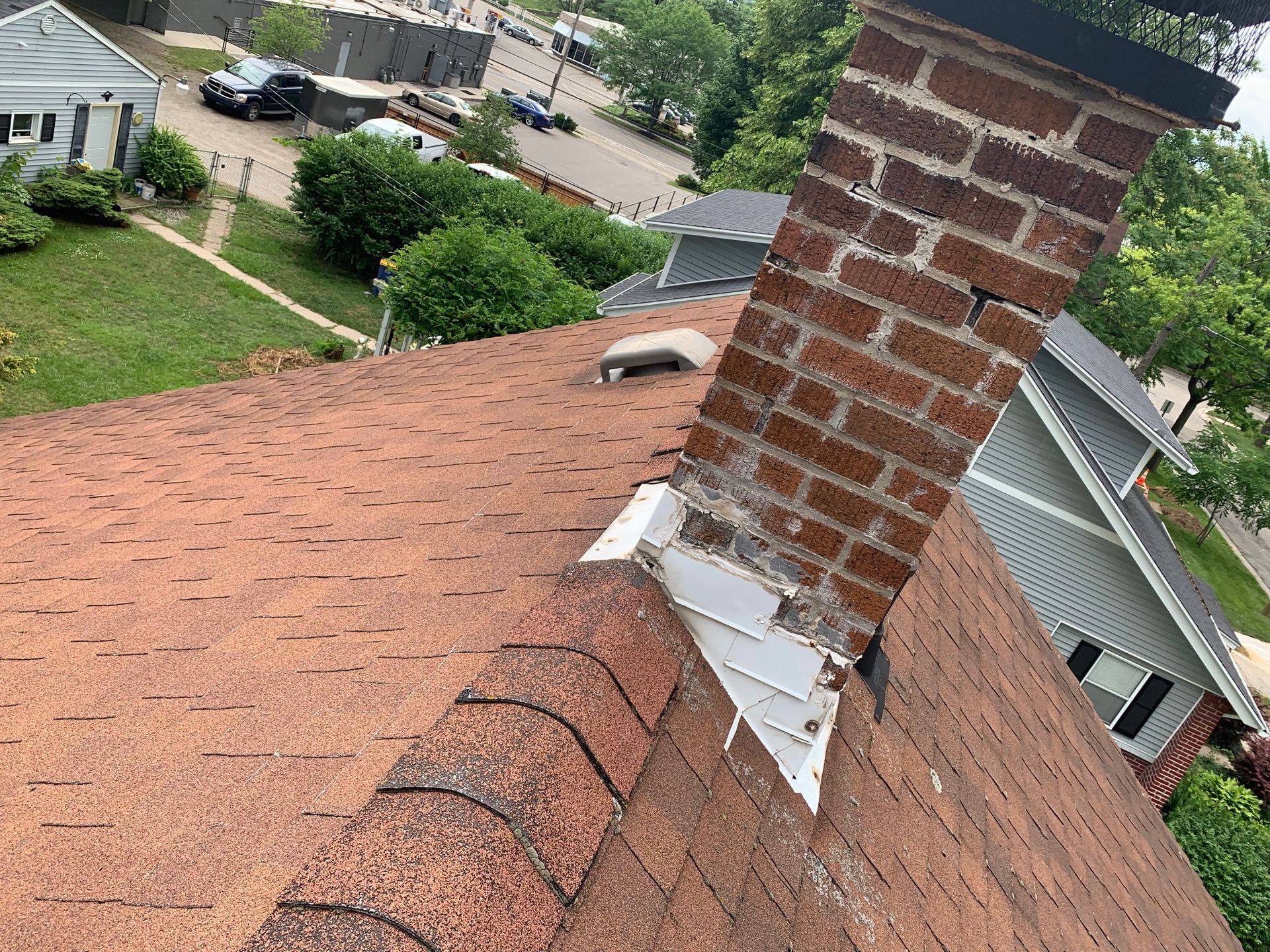 Steep shingle roof with brick chimney and flashing, viewed from above with neighborhood below.