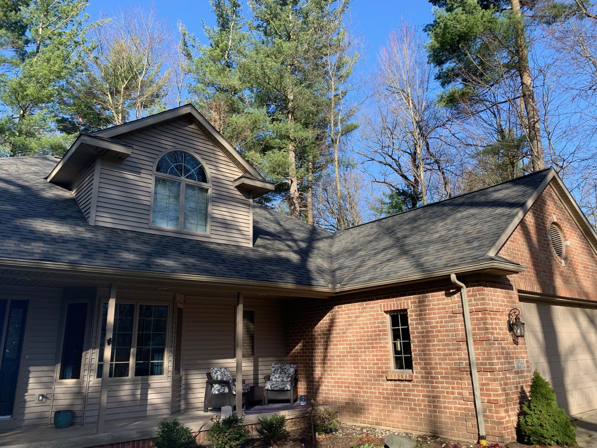 Brick-and-stone house with a front porch and tall pine trees under a clear blue sky