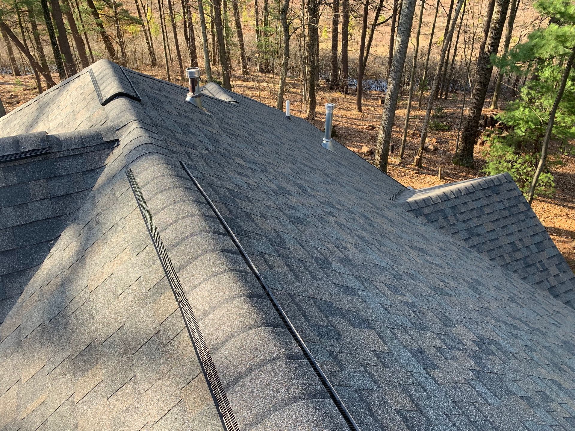 Gray shingled roof viewed from above beside a wooded area in autumn light