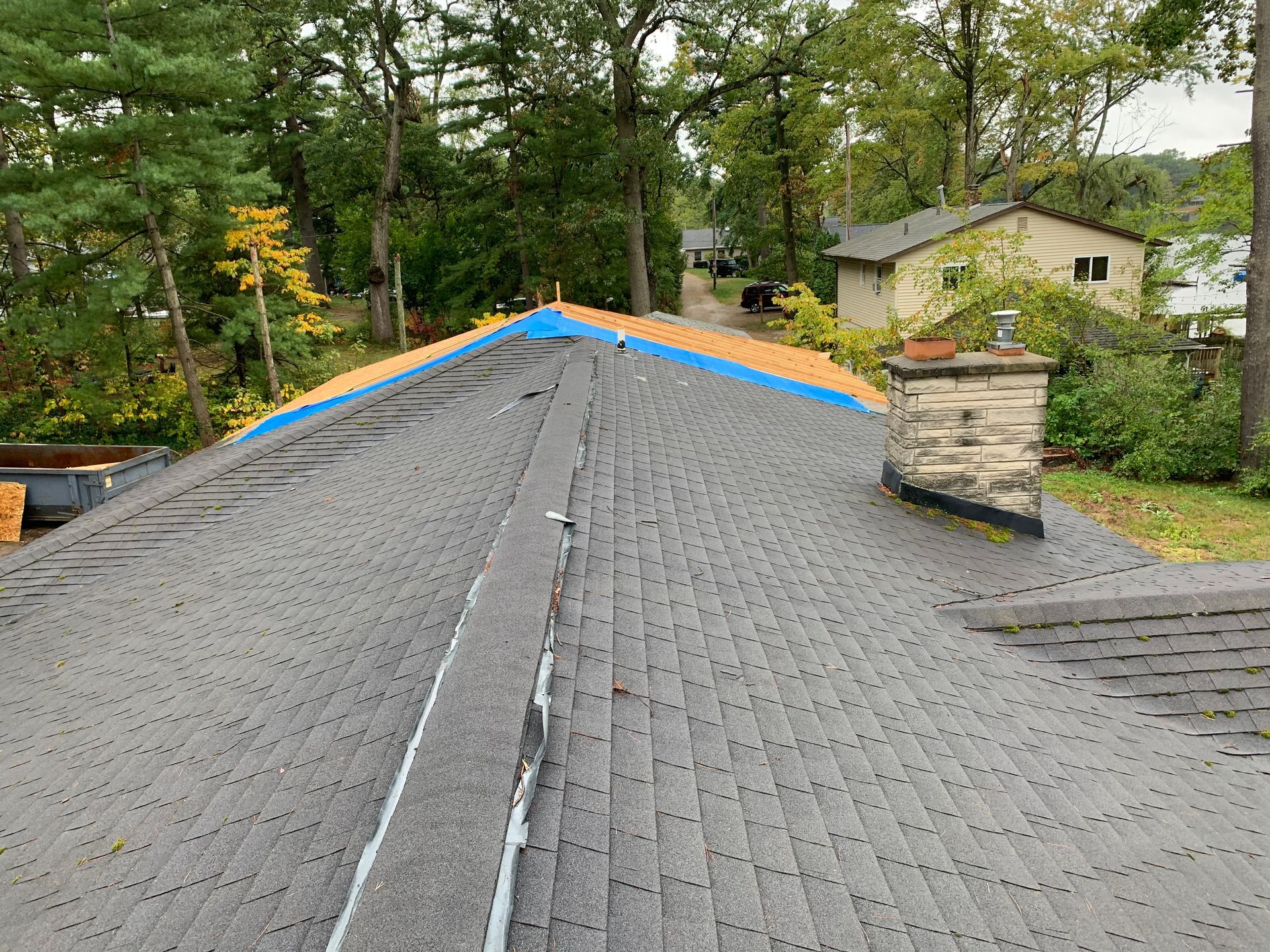 Roof with gray shingles under repair, blue tarp along the ridge, trees and a chimney nearby.