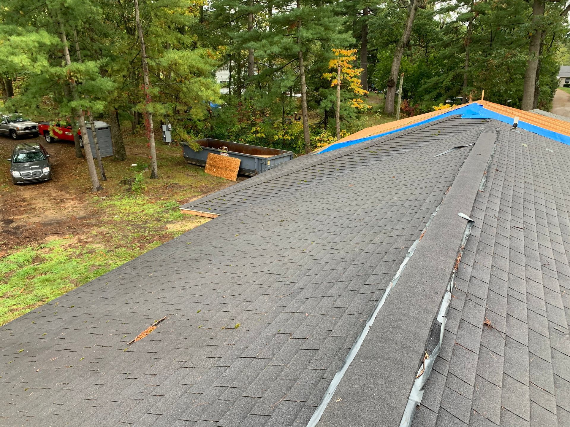 Roof shingles during replacement on a house in a wooded area, with blue tarp along the ridge.