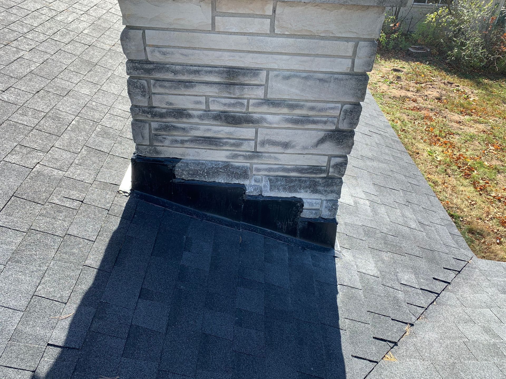 Black roof flashing around a stone chimney on a shingled roof with leaves nearby