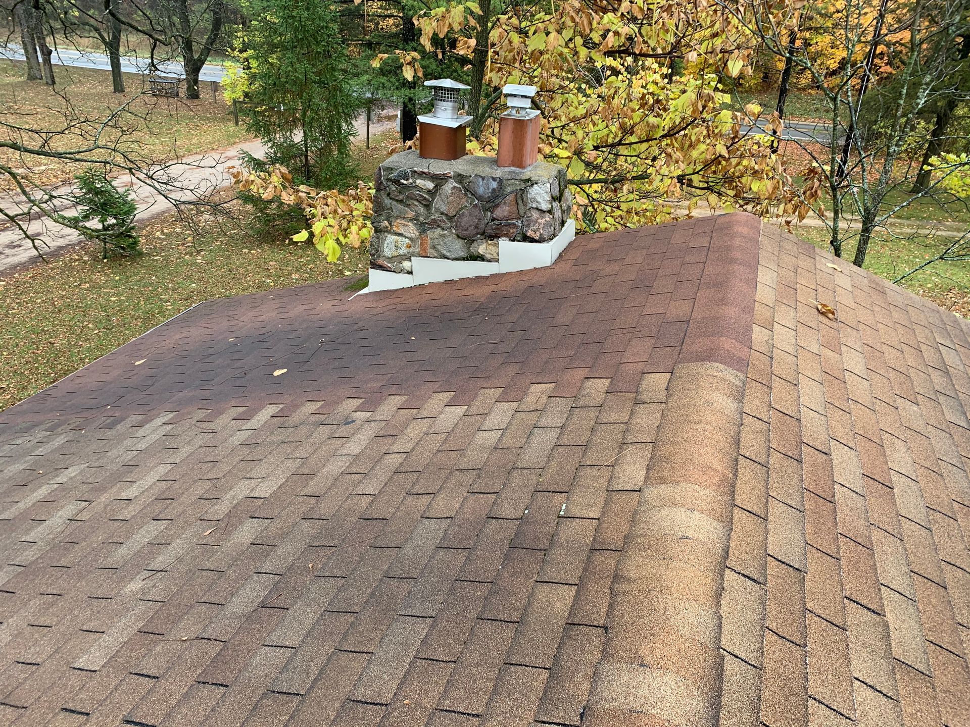 Brown shingled roof with stone chimney overlooking autumn trees and a path