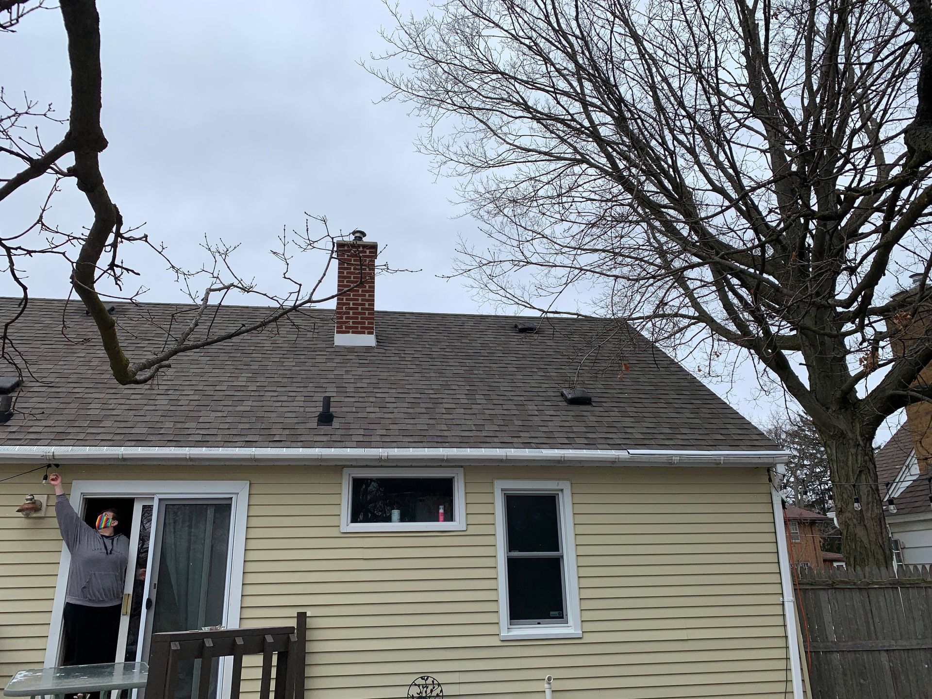 Person on a ladder painting the side of a beige house with a gray roof and bare trees.