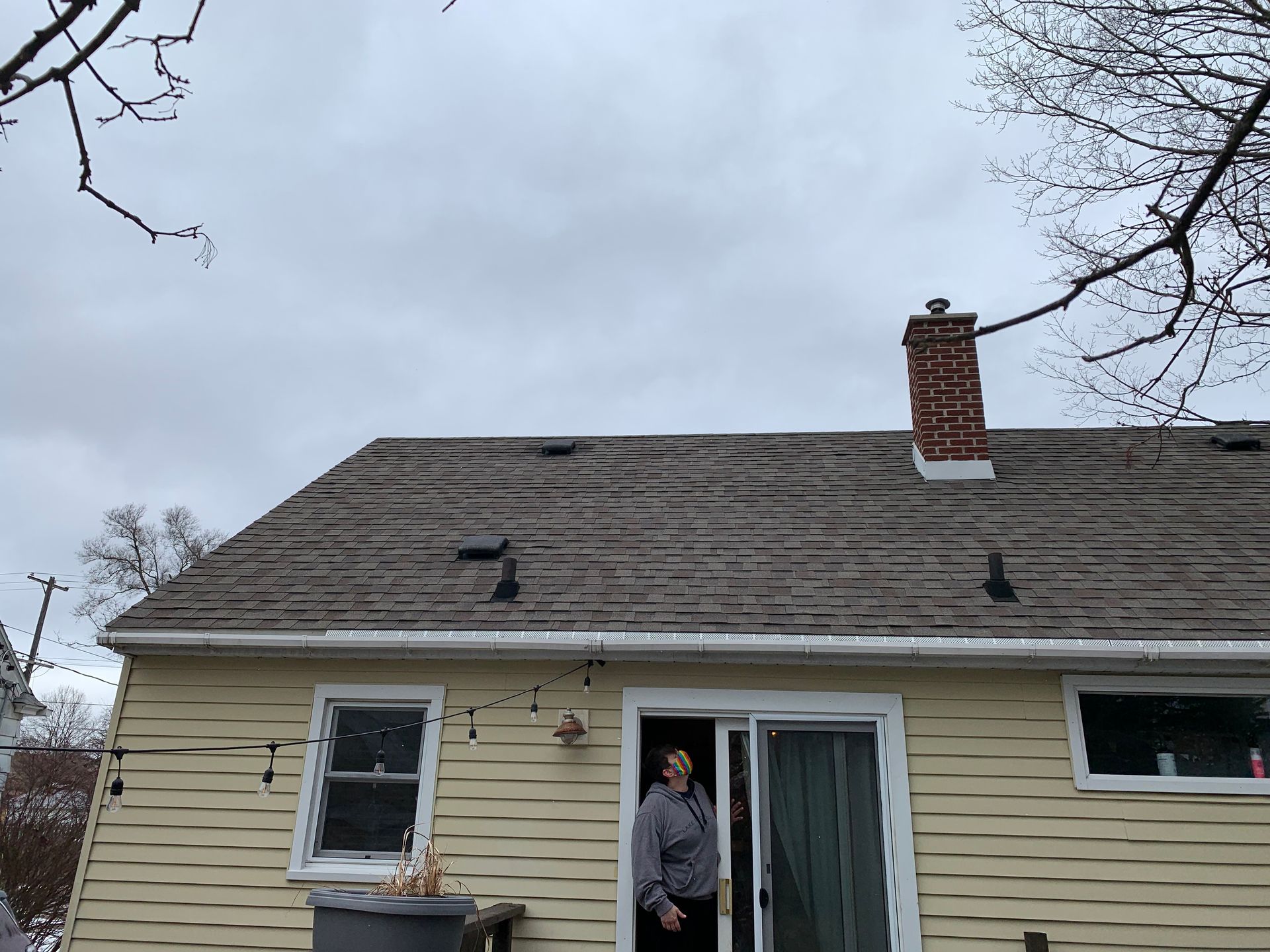 Person standing in a doorway of a beige house with a gray roof and brick chimney
