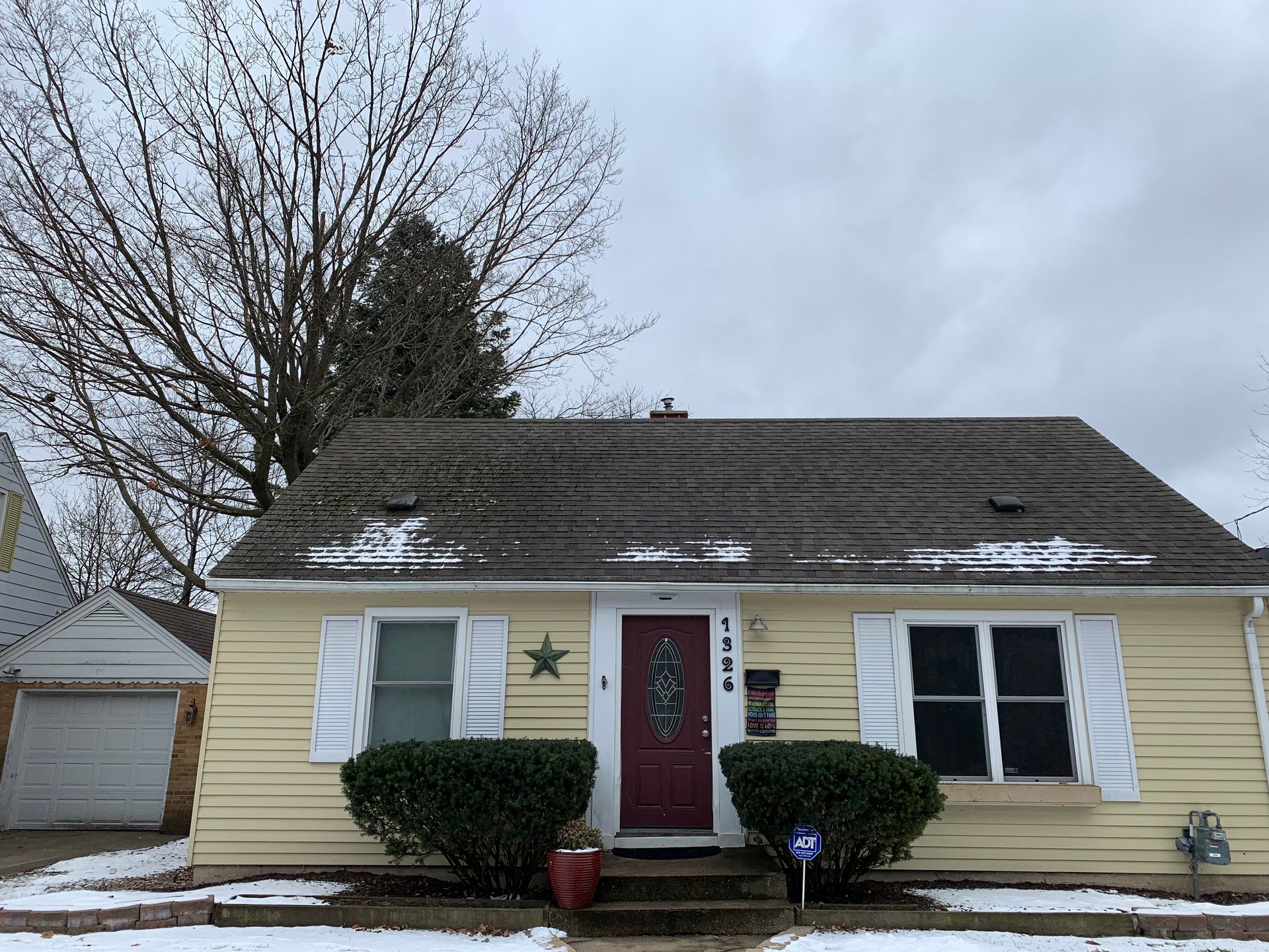 Snow-dusted yellow house with red front door, white shutters, and leafless trees under a gray sky