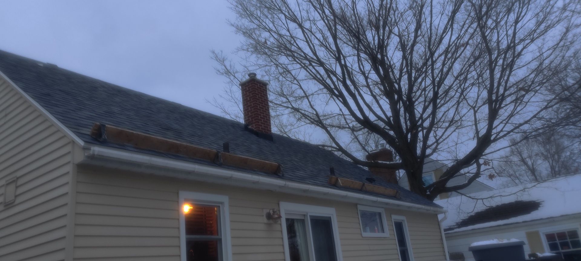 Snowy suburban house roof with a lit porch light and bare tree branches at dusk