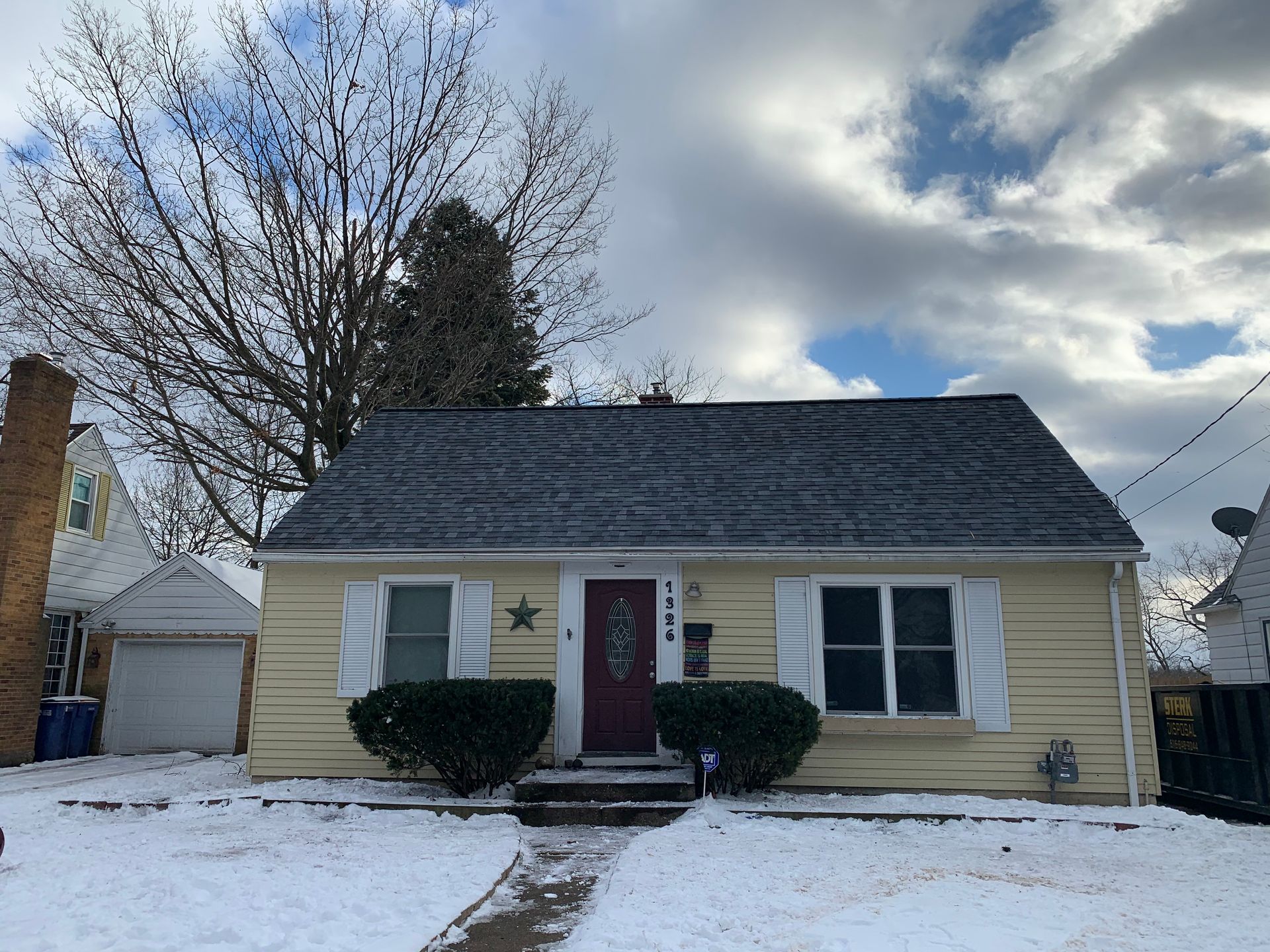 Snowy suburban house with beige siding, red front door, white shutters, and cloudy winter sky