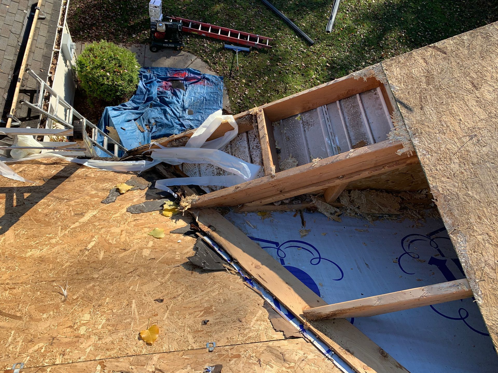 Damaged roof and attic opening with broken wood, blue tarp, and scattered debris outdoors.