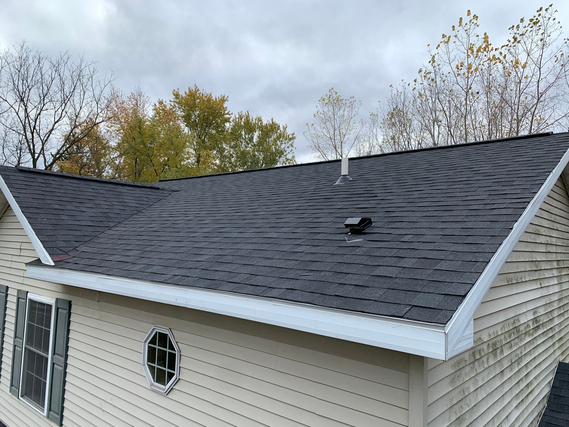 House roof with dark shingles, white trim, and a small roof vent against cloudy sky and autumn trees