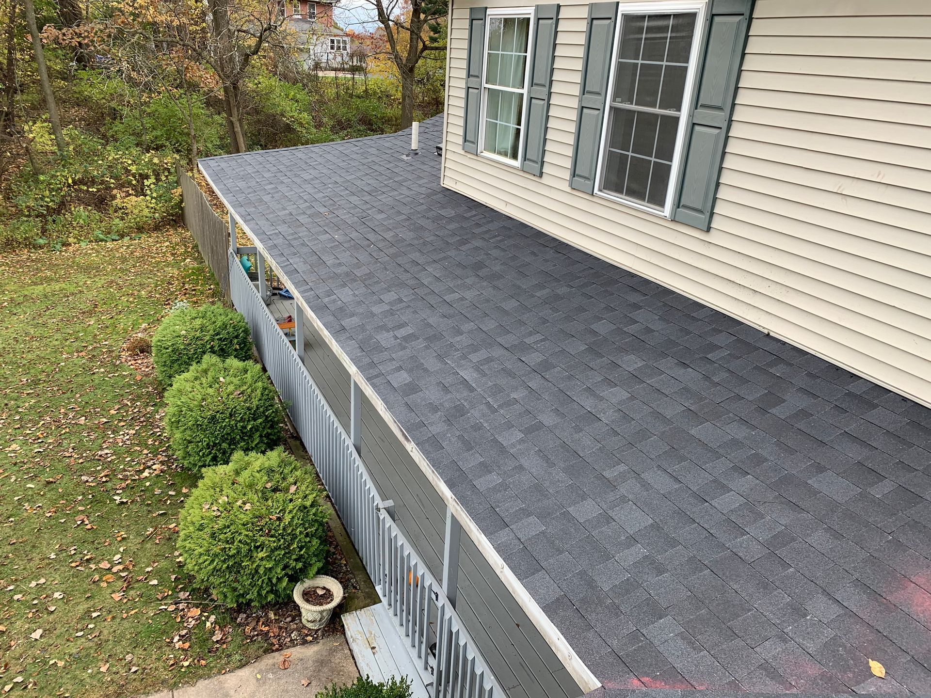 Roof with dark gray shingles over a porch beside beige house and trimmed bushes in a yard