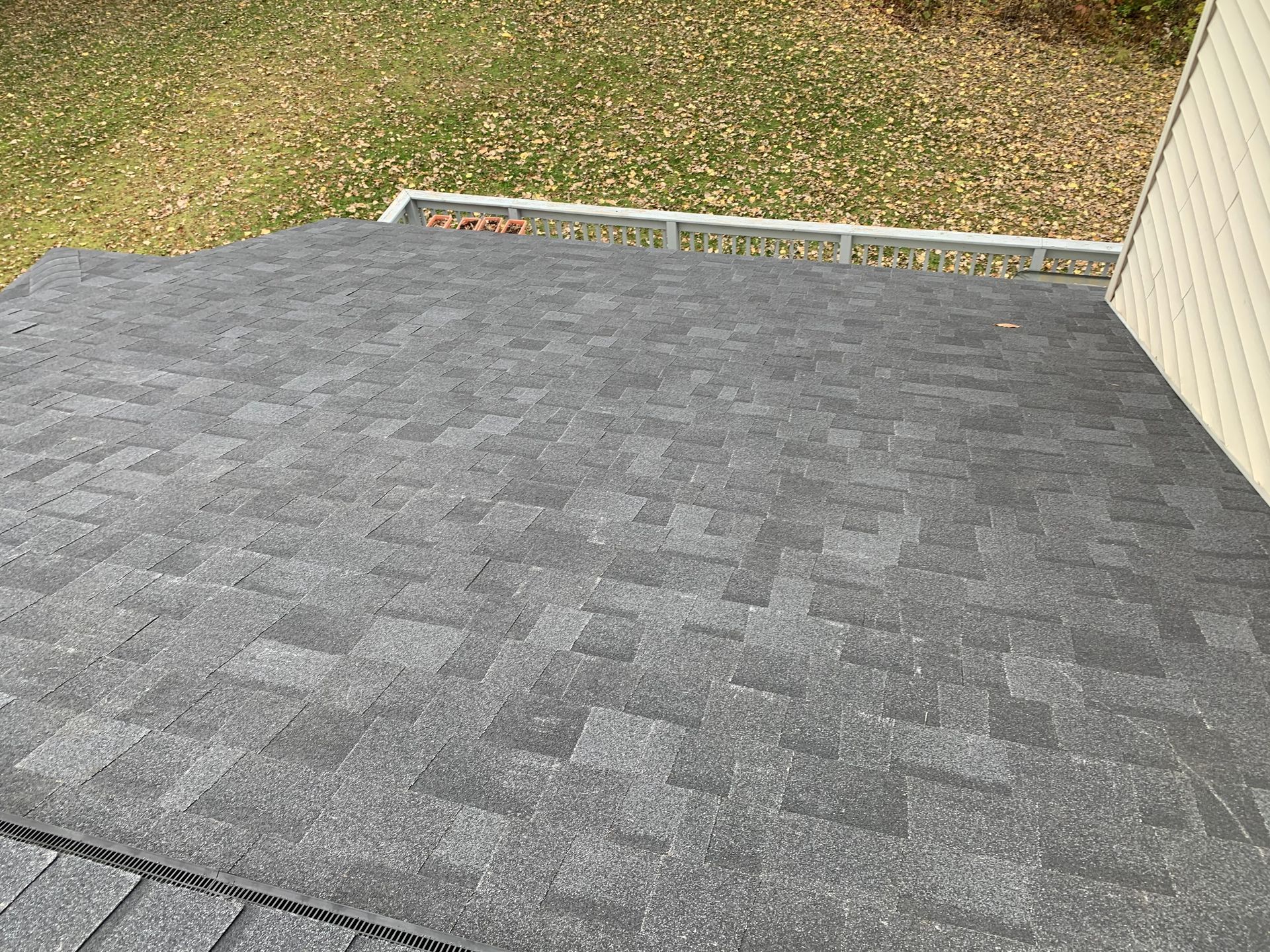 Gray shingled roof deck beside a house, with grass and a narrow gutter visible in the background